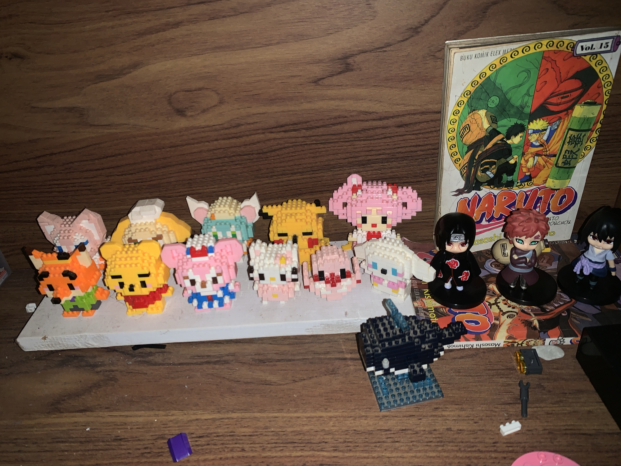 This image shows a collection of toys and other items on a wooden surface. In the background is a copy of "Naruto," Vol. 15, published by Elex Media Komputindo. In front of the book are three "Naruto" character figurines. There are also several small block-built figures including a fox, Pooh Bear, Hello Kitty, and other characters arranged on a white plank. Lego bricks are scattered on the right of the plank. The image appears to be taken indoors. The book is copyrighted by Masashi Kishimoto. The book says "Elex Media Komputindo," a publishing company headquartered in East Jakarta, Indonesia.