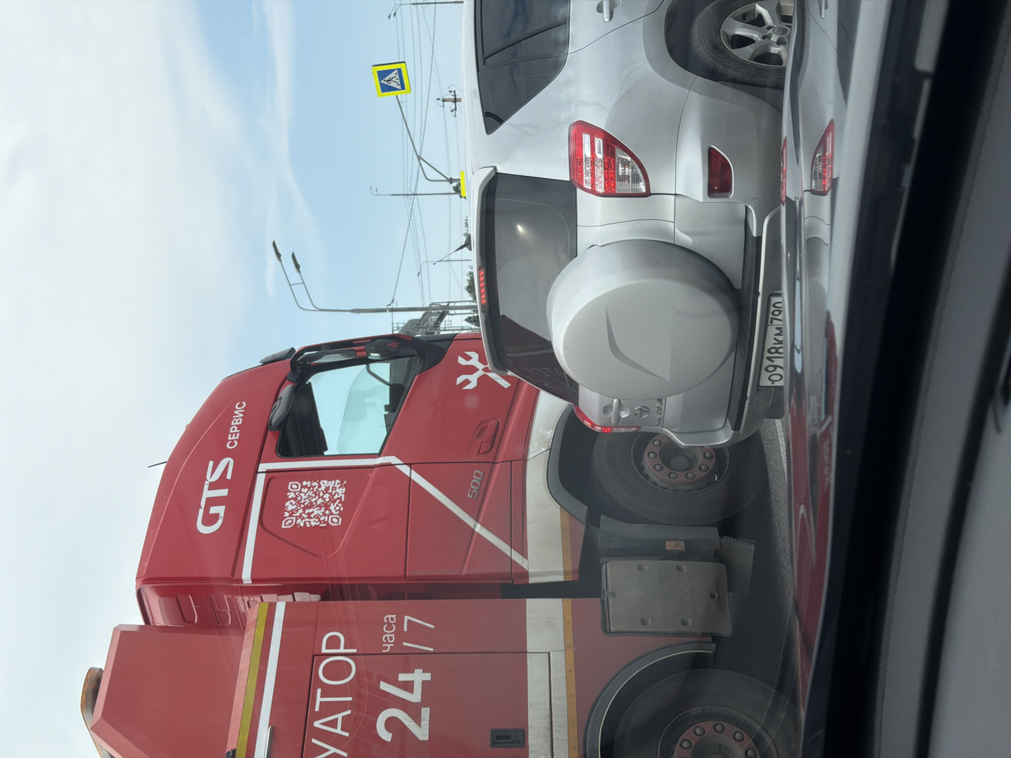 A red tow truck with "GTS СЕРВИС" and "УАTOR 24/7" written on its side is parked on a street. A silver SUV with a spare tire mounted on the back is behind the tow truck. The license plate of the SUV is visible and reads "0918KM 790". There is a pedestrian crossing sign in the background. The scene appears to be a street in the city of Rodnik, Russia. The weather is overcast and the sky is visible with clouds. The lighting suggests it is daytime.