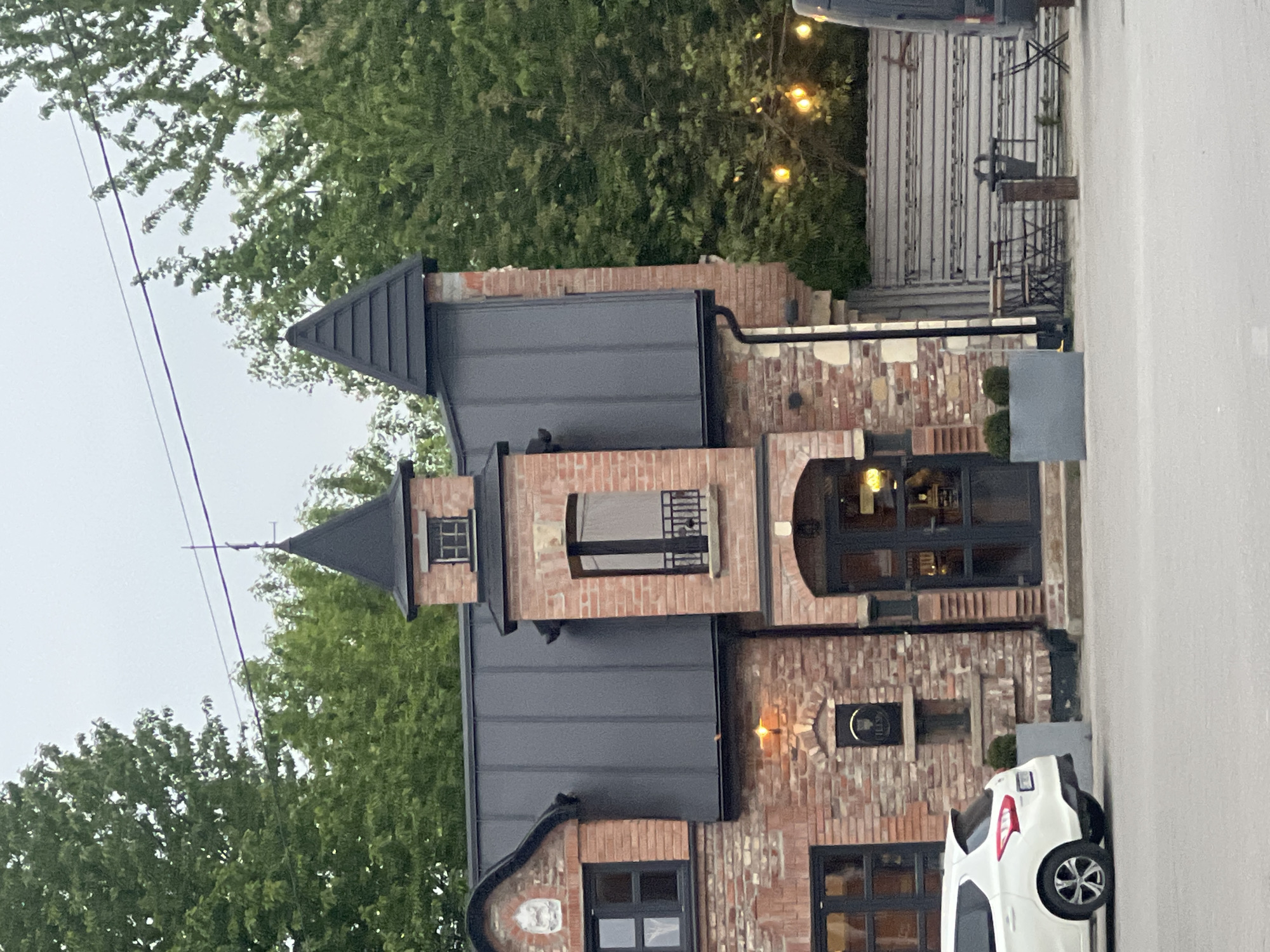 The image depicts a brick building with a distinctive turret-like structure and a modern, dark-colored roof. The building has multiple windows and what appears to be an arched doorway. There is a white car parked on the right side of the image, partially obscuring the building's facade. Green trees surround the building, and there are overhead wires visible in the sky. The location is Horodok, Ukraine.