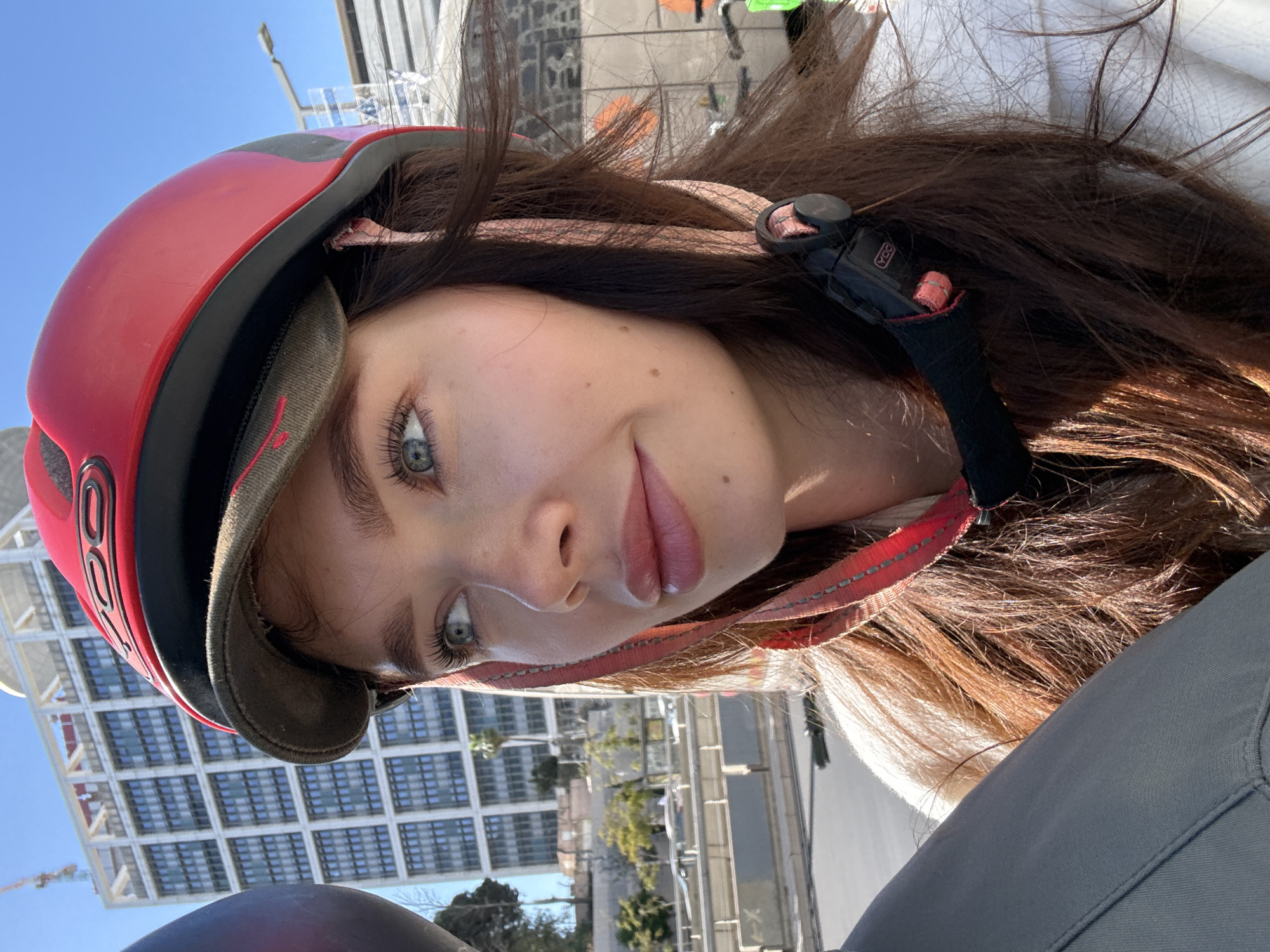 A young woman is captured in a sunlit outdoor selfie, her head tilted slightly, wearing a red and black helmet over a brimmed cap. Her long, dark brown hair flows around her shoulders. She has striking blue-green eyes and her lips are slightly parted in a subtle smile.
The helmet features a visible white "POC" logo on the side, indicating the brand. A black chin strap with a red patterned strip is visible, secured by an adjustment clip on the left side of her face. The material of her outfit includes a white top and a greyish-green fabric, possibly pants, visible on her lower right.
The background reveals a clear, bright blue sky and modern urban architecture. Tall buildings with a grid-like window pattern dominate the backdrop, suggesting a bustling city environment consistent with Tel-Aviv, Israel. Distant structures and a street light are also discernible under the bright daylight. The overall impression is one of activity in an urban setting, likely for a sport or commute given the helmet.