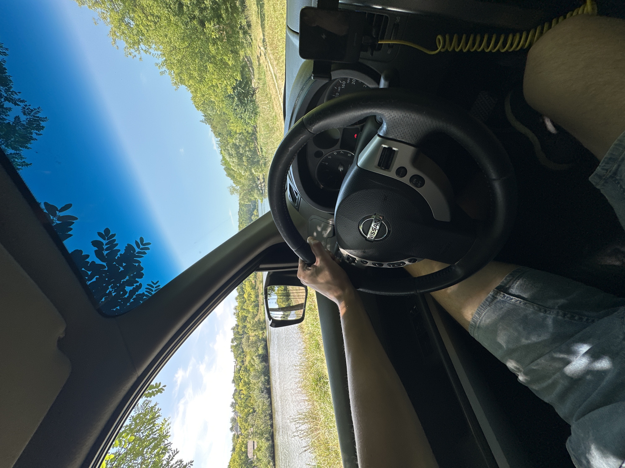 An interior shot from the driver's perspective of a Nissan vehicle, taken on a bright, sunny day. The driver's left arm and hand, wearing denim shorts, are visible, gripping the steering wheel. The dashboard, including the Nissan logo on the steering wheel and a phone mount with a charging cable, is in focus. Outside the vehicle, through the open window and windshield, a tranquil scene unfolds. A calm body of water, likely a river or lake, is visible to the left, with green, tree-covered hills rising in the background. The reflection of trees and sky can be seen on the water's surface. The overall environment suggests a rural or semi-rural setting, possibly on the outskirts of Makiv, Ukraine, given the natural landscape. The sun is high in the sky, casting shadows and illuminating the scene.