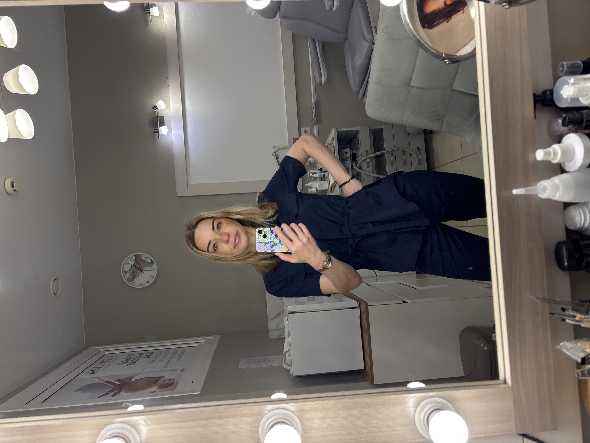 A woman is standing in front of a mirror, taking a selfie with her phone. She is wearing a dark blue jumpsuit with a belt. In the background, there is a medical chair and some equipment, suggesting that she is in a dental or medical office. There is also a clock on the wall and a poster on the bottom left. There is a small round mirror attached to the side of the main mirror. The location is in the city of Unknown, Unknown.