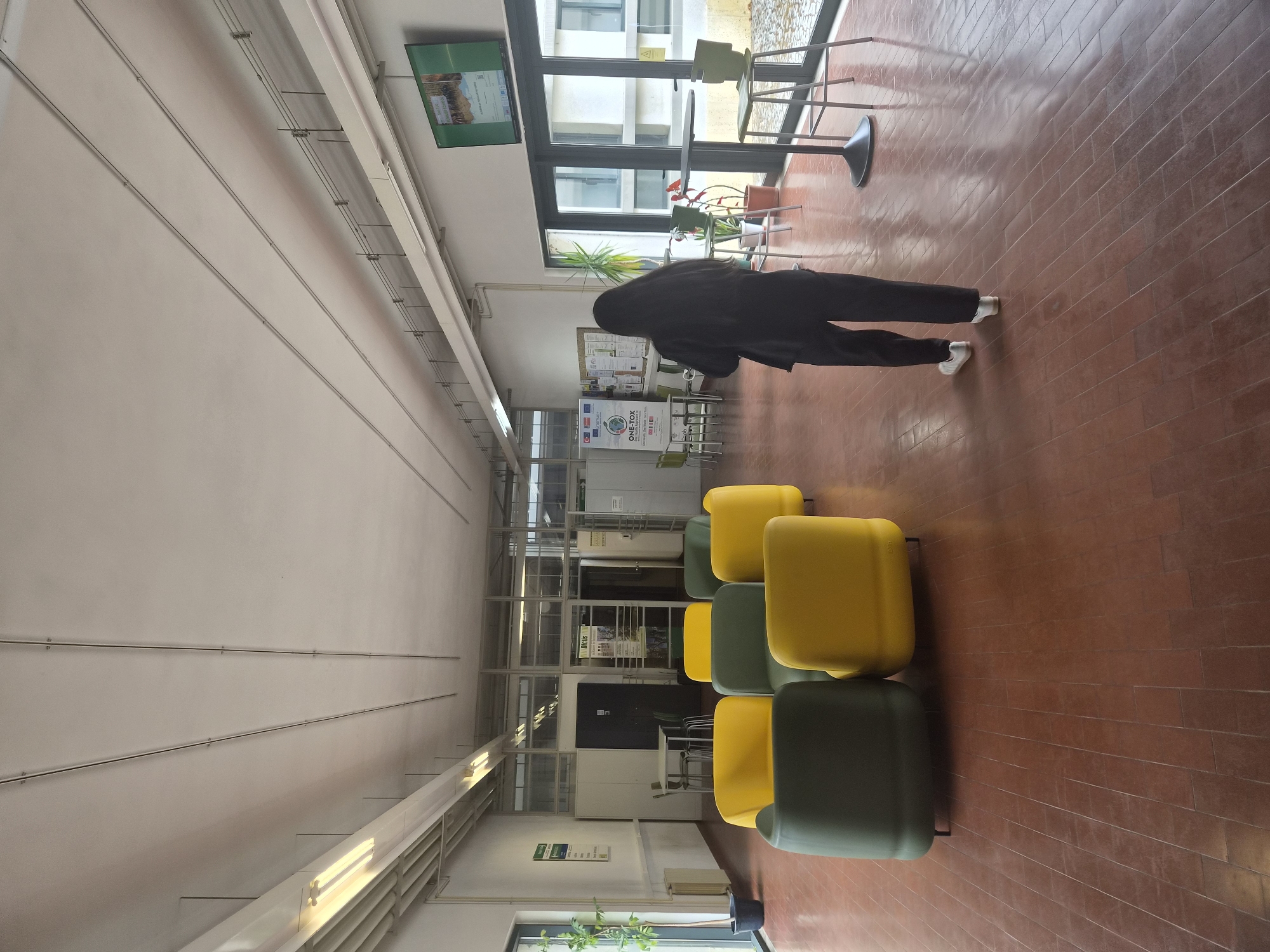 The image depicts a wide, indoor hallway with a reddish-brown tiled floor. The hallway is illuminated by fluorescent lights running along the ceiling. On the left side, there are large windows and a plant in a black pot. A section of the hallway features a cluster of brightly colored, modern armchairs, some in yellow and some in dark green. Further down the hallway, there are tables and chairs, and glass-partitioned rooms. A large screen displaying an image is mounted on the wall above a set of glass doors on the right. A person with long black hair, wearing a black top and pants, is walking away from the camera towards the right side of the hallway. The environment suggests a public space, possibly a university or office building. This scene is located in the campus of the Polytechnic Institute of Bragança (IPB), in the city of Bragança, Portugal.