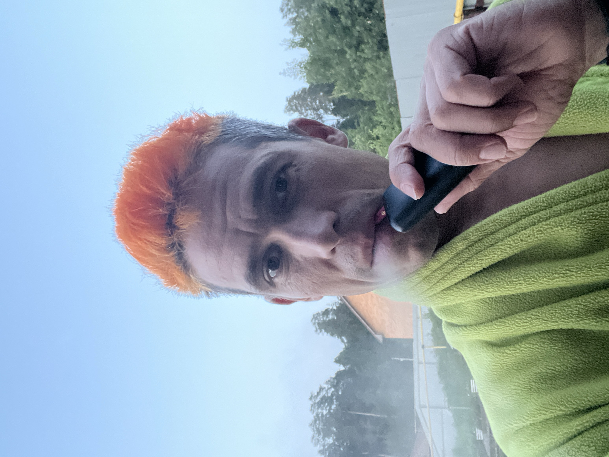 A close-up selfie shows a person with bright orange hair and a green robe. They are holding a black electronic cigarette to their lips, with a faint plume of vapor visible. The background features a hazy sky, trees, and a glimpse of a building. The setting appears to be outdoors, possibly a backyard or a park, given the presence of trees and a fence. The overall ambiance suggests a casual, relaxed moment. The location is identified as Chaikovsky, Russia.