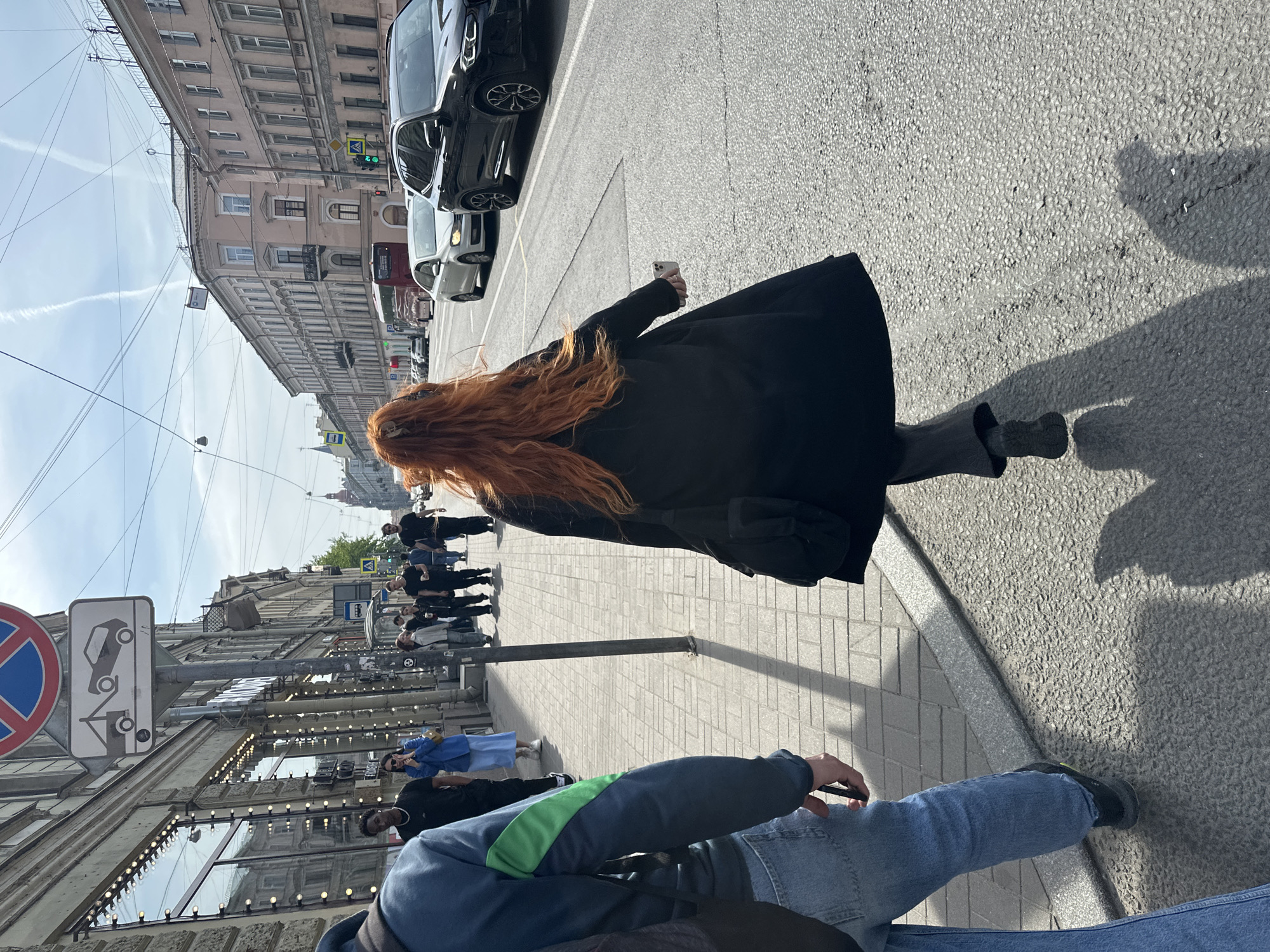 A street-level photograph shows a pedestrian scene in St. Petersburg, Russia. The image is oriented with the sidewalk on the left and the roadway on the right. In the foreground, a person is sitting, wearing jeans and a blue jacket with a green stripe. In the middle ground, a red-haired person in a long black coat walks away from the viewer, holding a mobile phone in their right hand. A line of people walks along the sidewalk in the same direction as the red-haired person. Street signs are visible, indicating traffic regulations. Cars are parked on the right side of the road. Buildings with multiple stories line the left and right sides of the street, and a pale blue sky with contrails is above. The pavement shows patches of shadow.