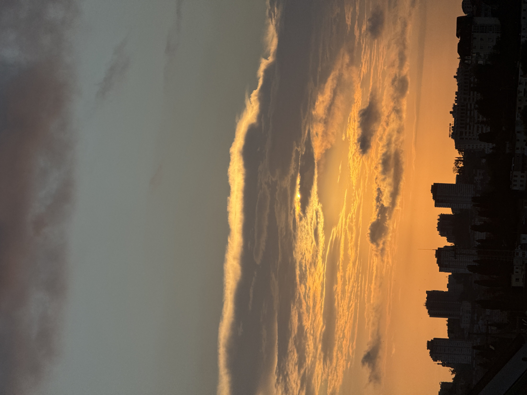This is a medium shot, eye-level, horizontal photograph of a cityscape at sunset. The sky is filled with clouds, illuminated by the setting sun, casting an orange glow across the scene. In the foreground, a silhouetted cityscape with numerous buildings, including apartment blocks and some smaller structures, stretches across the horizon. The buildings are densely packed, and trees are visible in silhouette between and in front of them. The overall atmosphere is serene, with the warm colors of the sunset dominating the sky. The location is the city of Sochi, Russia.