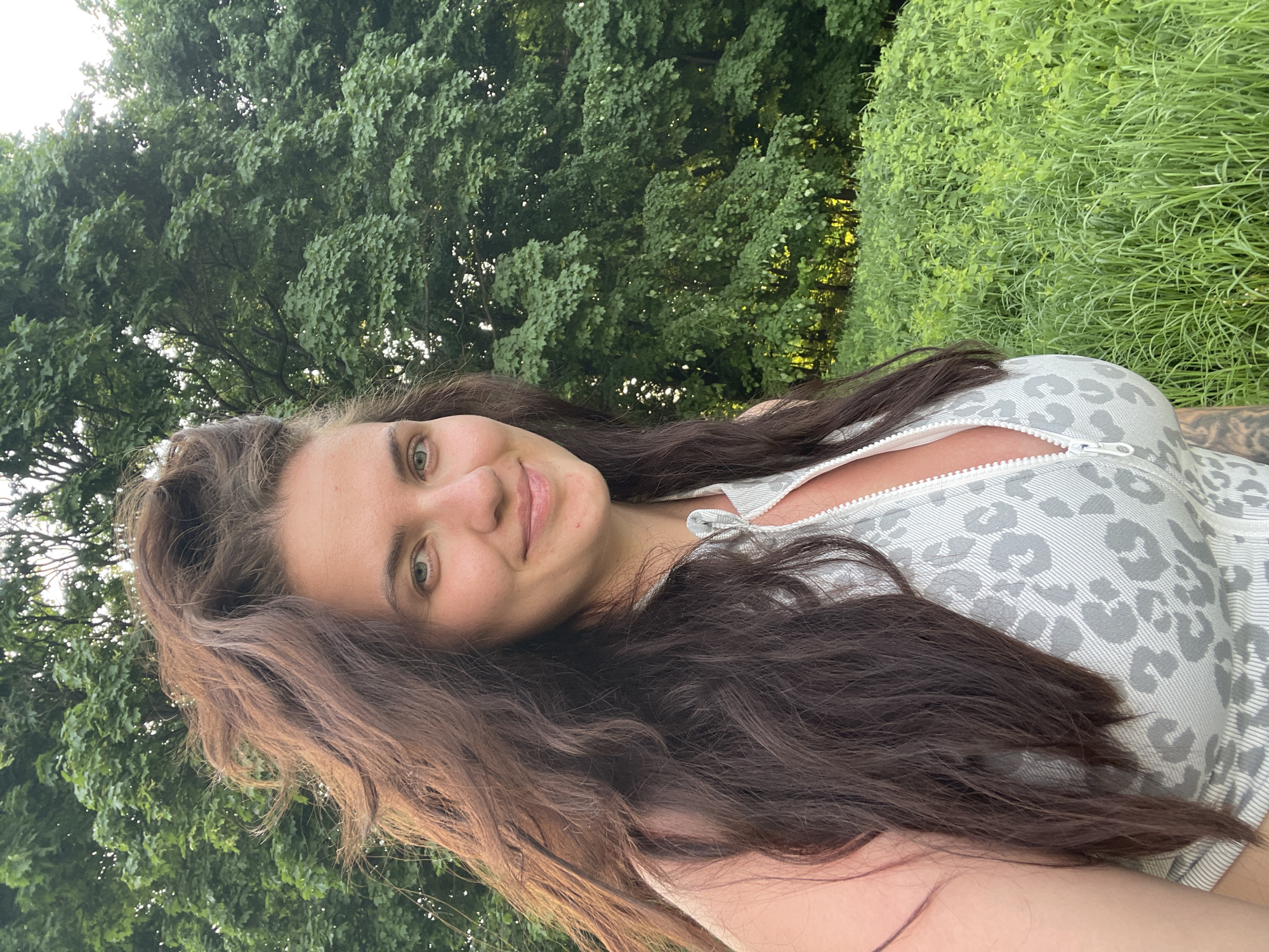 The image is a selfie of a woman with long, dark brown hair. She is smiling slightly and looking at the camera. She is wearing a light gray top with a leopard print pattern and a zipper. The background consists of lush green trees and tall grass, suggesting an outdoor location. There are no indications of a specific location in the city of Ostrov, Russia.