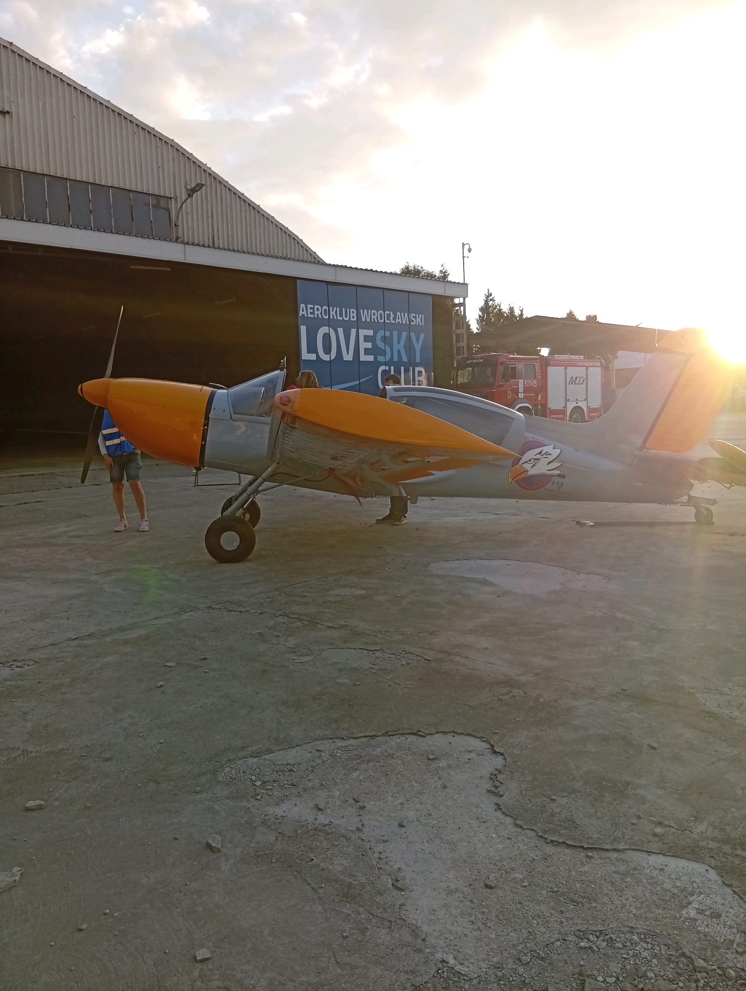 The image depicts a small, propeller-driven aircraft parked on a concrete tarmac outside a hangar. The aircraft, primarily silver with orange accents on the wings and nose, features an eagle emblem on its fuselage. Behind the aircraft, two red fire trucks are visible, one with the Polish inscription "OSP POZOROWICE" and the number "577435," and another with "MD" on its side. The hangar has a large open entrance and signage that reads "AEROKLUB WROCŁAWSKI" and "LOVESKY CLUB." The setting appears to be an airfield, possibly associated with an aeroclub, in Szewce, Poland, given the visible inscriptions. The sun is setting, casting a warm glow and long shadows across the scene. Two individuals are partially visible near the aircraft. One, wearing a blue and white vest and shorts, stands by the front of the plane. Another person is seated in the cockpit. The ground is a mix of cracked and weathered concrete, with scattered small stones. The sky is cloudy, with patches of bright sunlight breaking through.