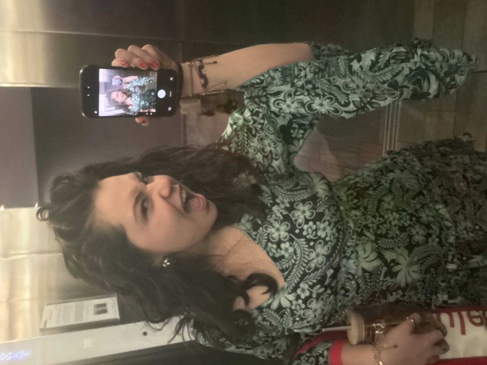 A woman with dark hair is holding up a smartphone, taking a selfie in an elevator. She is wearing a dress with a dark floral pattern on a light green background. She has red nail polish and is wearing jewelry on her wrists. The elevator has a metal interior.