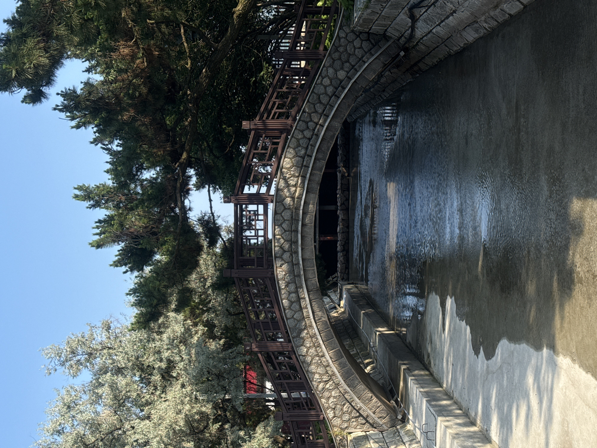 A stone arch bridge with wooden railings spans a narrow canal. The canal has concrete banks and is filled with dark water, reflecting the trees and sky above. The bridge is decorated with stone and wood, and the railings have a lattice pattern. Trees with green and silver foliage line the banks of the canal, with more dark green trees visible in the background. The sky is clear and blue. The image appears to be taken during daylight hours. The location is Beidaihe District, Qinhuangdao, China.