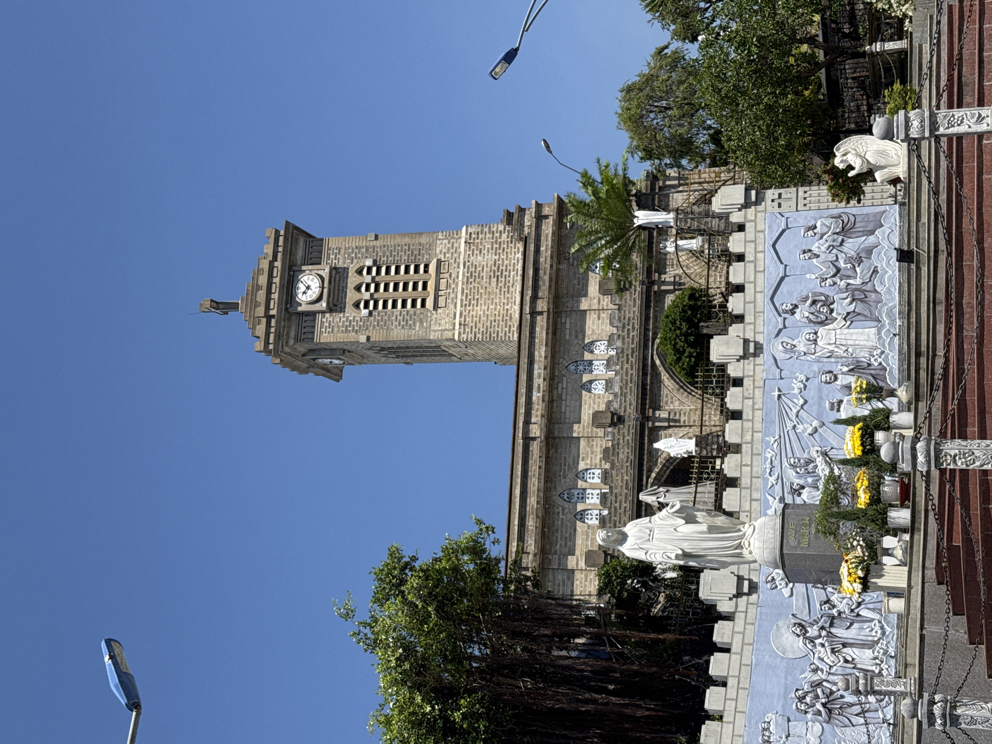This is a street-level, medium shot of the Nha Trang Cathedral, located in the Nha Trang Ward of Vietnam. The cathedral tower is the dominant feature, built of grey stone with a clock near the top. The clock face shows the time is around 10:10. The tower is multi-tiered, and a cross is perched at the top. The main structure of the cathedral is also built from the same grey stone.
To the right of the cathedral structure, along what appears to be a walkway, there are multiple statues and relief panels depicting religious scenes. The statues are white and appear to be the Virgin Mary. Floral arrangements are placed near the statues. The relief panels are light blue with white figures. A chain railing is visible in the foreground, delineating the boundary of the walkway or stairs.
Trees are visible around the cathedral, suggesting that it is surrounded by green space. The sky is clear and blue. Blue streetlights are on tall poles and present. The overall impression is one of a well-maintained religious building in a sunny environment.