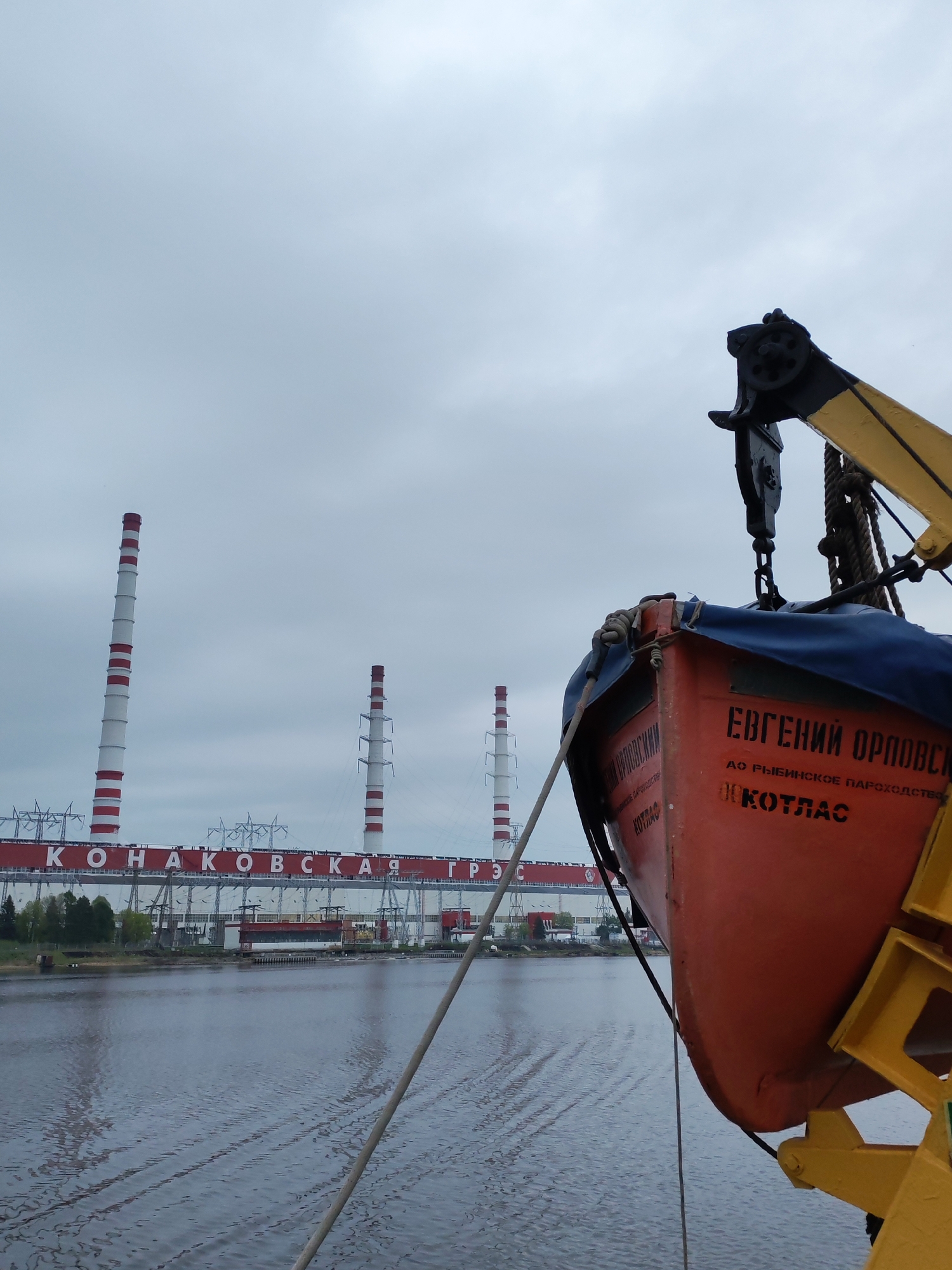 An eye-level outdoor shot reveals an orange lifeboat marked with Cyrillic script hanging from a yellow davit, with a thermal power plant in the background under an overcast sky. The plant, identified as "KONAKOVSKAYA GRES", features four tall white chimneys with red horizontal stripes. The plant building, with a red stripe along the top, runs horizontally across the frame behind a body of water, likely a river or large lake, with visible ripples. The lifeboat is suspended by thick ropes and a black metal mechanism. The water’s surface appears calm with faint ripples, possibly indicating a slight breeze.