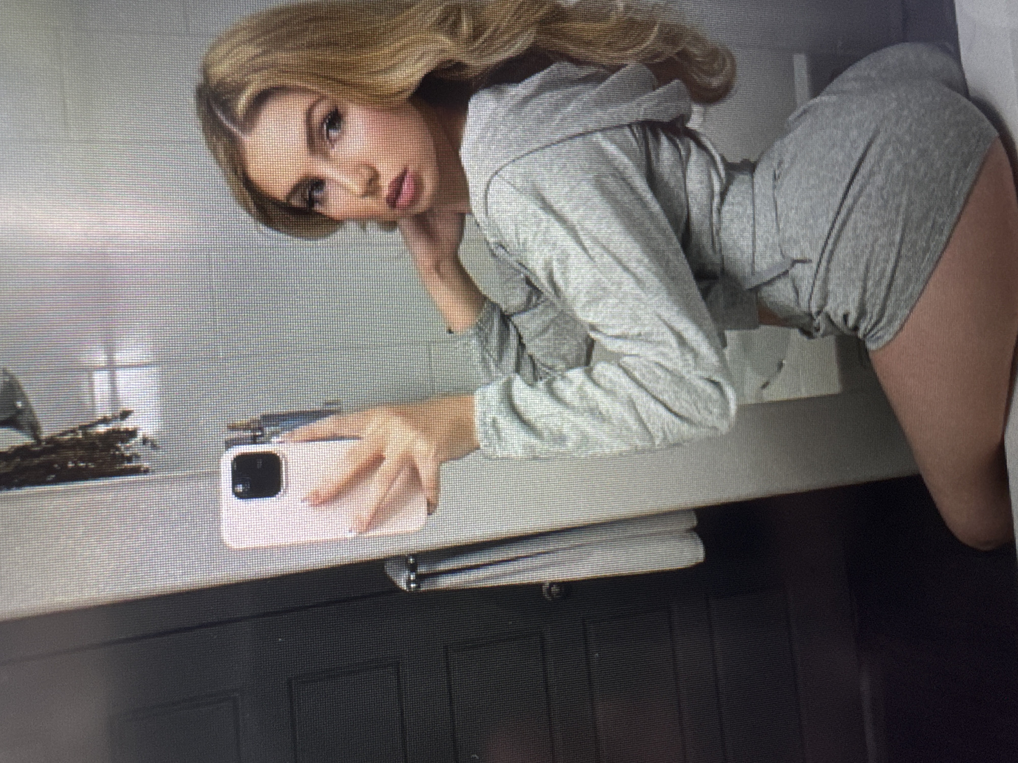 The image displays a young woman posing indoors, likely in a bathroom or vanity area. She has long, wavy blonde hair and is wearing a full face of makeup, including winged eyeliner and reddish-pink lipstick, with a confident, direct gaze and a slight pout.
She is dressed in a grey two-piece outfit comprising a cropped hoodie and form-fitting high-waisted shorts. Her body is angled, leaning over a light-colored counter. Her left hand, with painted nails, rests on the counter, holding a light-colored smartphone. Dark, decorative items are visible on the counter to the left of the phone.
The background behind her features a light grey wall or mirrored surface. Below the counter, a dark, paneled cabinet or vanity unit is visible, with a folded white towel or fabric on a rack underneath.
The photograph captures a posed moment, possibly a mirror selfie. The image itself appears to be a photograph taken of a digital display, evidenced by the visible pixel grid pattern and slight blurring. No specific time of day or weather can be determined from the content. There is no visible text in the image, and no visual cues confirm the location as Varna, Bulgaria.