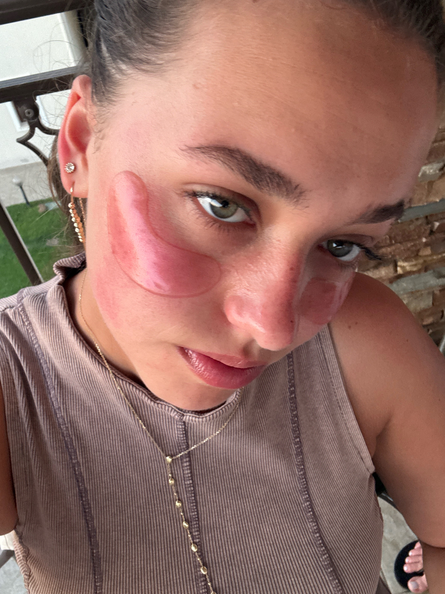 A woman is captured in a close-up, downward-facing selfie, taken outdoors. She is wearing a taupe ribbed tank top and a delicate gold necklace. Under her eyes, she has applied two large, translucent pink under-eye patches. Her hair is pulled back, revealing a prominent ear with a stud earring and a dangling chain earring. The background suggests a balcony or patio setting with a stone wall and a glimpse of greenery. The lighting indicates it is daytime, likely late afternoon given the warm tones. The image does not contain any identifiable landmarks or features that would allow for precise location within Nea Flogita, Greece.
