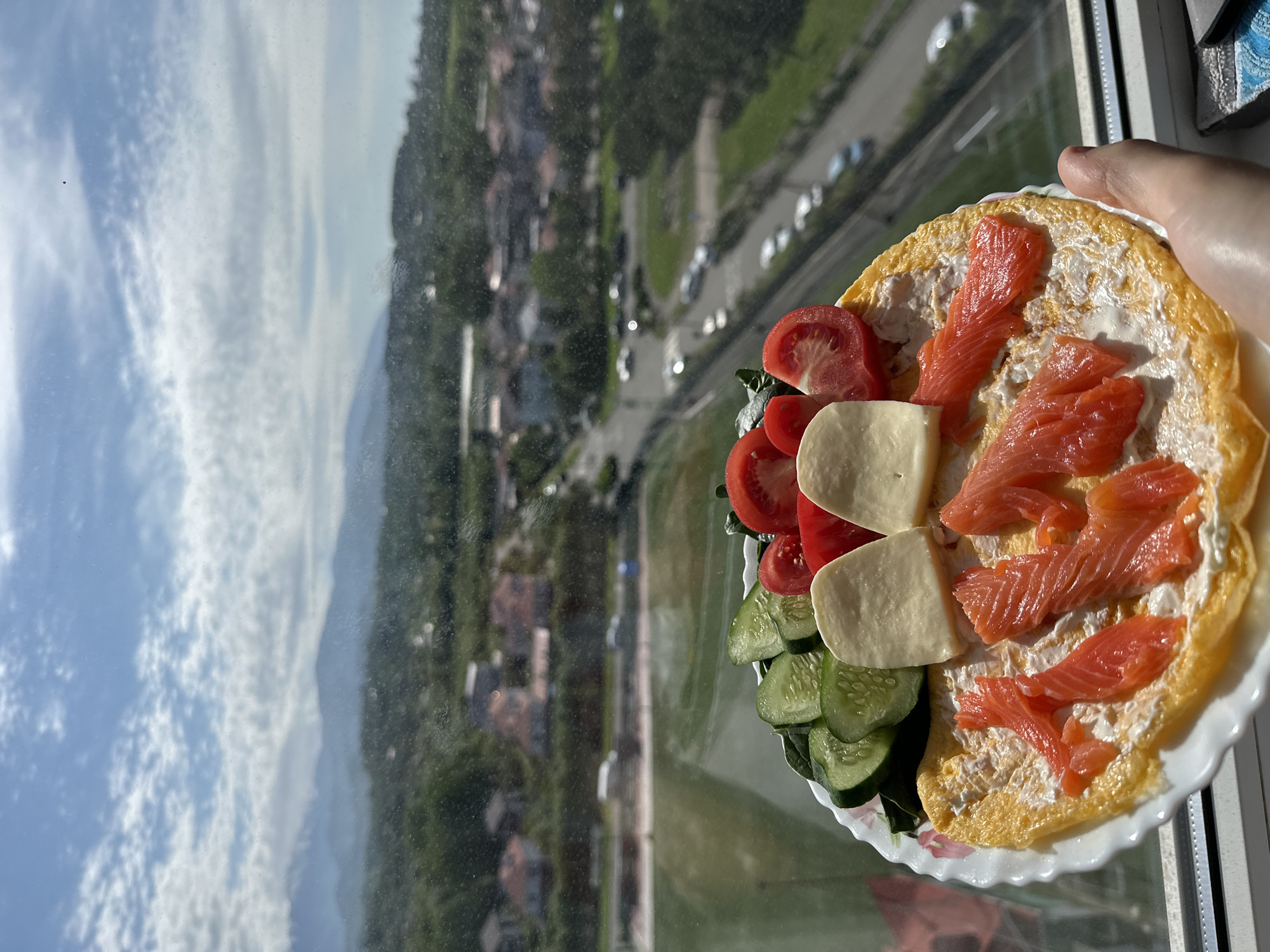 A person holds a plate containing an omelet with cream cheese, slices of smoked salmon, sliced tomatoes, pieces of white cheese, and sliced cucumbers. The plate is held in front of a window with a view of a cityscape, mountains, and the sky. The cityscape appears to be Sochi, Russia, due to the mountainous terrain, buildings, and road infrastructure visible. The sky is partly cloudy.