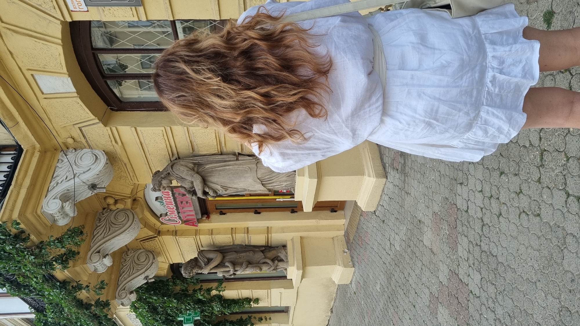 The image shows the facade of a building in Chernivtsi, Ukraine, featuring yellow stucco walls, ornate architectural details, and statues. A woman with long, wavy brown hair, viewed from behind, is standing on a paved walkway in front of the building. She is wearing a white linen dress with a cinched waist and ruffled hem, and a white bag is slung over her shoulder. The building appears to be an older establishment, possibly a pharmacy, with a sign above the entrance that reads "ЄЛИНА АПТЕКА" (Yelina Apteka). Green ivy grows on the upper part of the building. The paving stones on the walkway are a mix of grey and reddish-pink interlocking tiles.