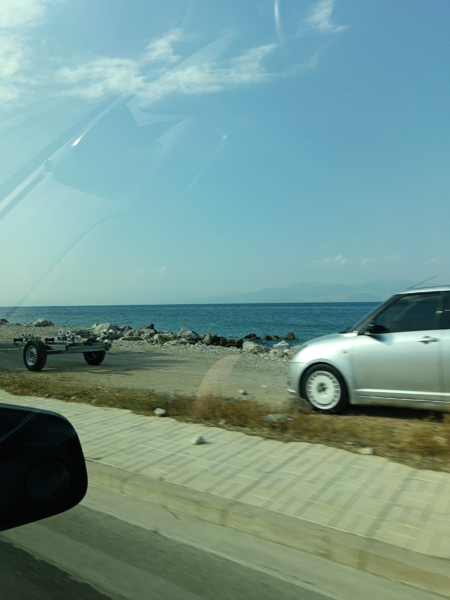 The image captures a scene from within a vehicle traveling along a coastal road. The foreground shows a portion of the vehicle's interior, including a side mirror and a reflection of the sky and clouds on the windshield.
The exterior scene reveals a clear blue sky with scattered clouds. The sea stretches out to the horizon, meeting a distant mountain range. The coastline is rocky, with a mix of sand and pebbles. In the mid-ground, a silver car is driving on the road. To the left of the car, a boat trailer is visible on the side of the road. The road itself is paved with interlocking bricks, and there is a strip of dry grass and vegetation bordering it. The overall impression is a scenic drive along the coast of the Municipal Unit of Sikyona, Greece.