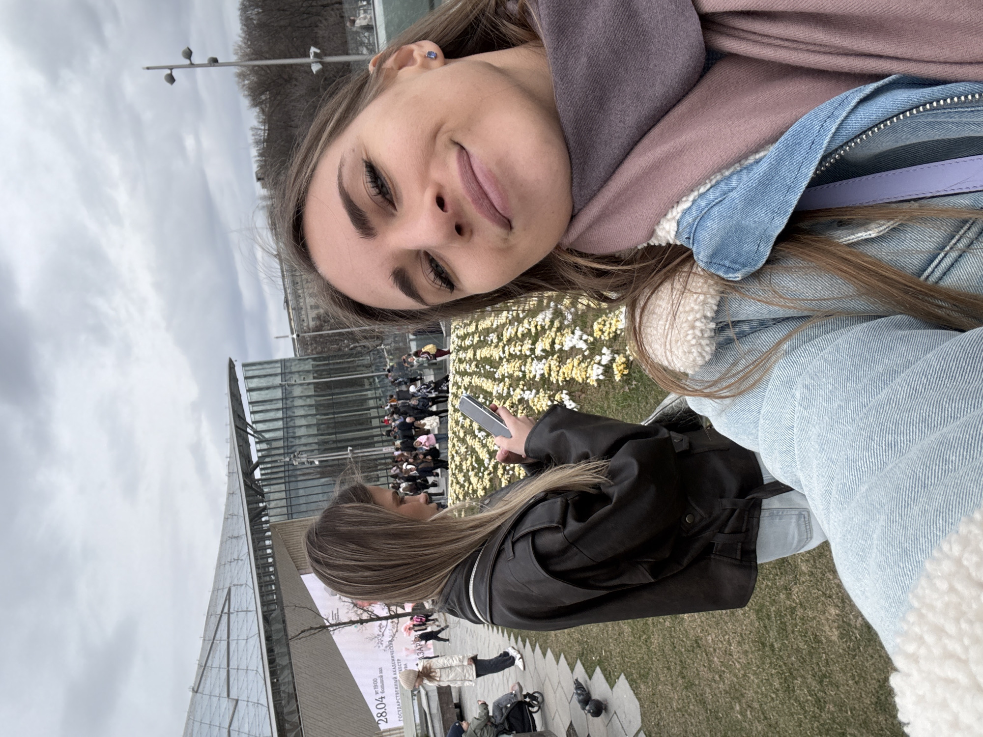 A selfie-style image captures two young women outdoors on an overcast day in Zaryadye Park, Moscow, Russia.
In the foreground, a woman with long brown hair, wearing a light blue denim jacket with white sherpa lining and a purple scarf, holds the camera, looking slightly upwards with a faint smile. A blue stud earring is visible on her left ear.
Behind her and to the left, another woman with long brown hair, wearing a dark brown leather-like jacket and light blue jeans, stands with her back partially turned, engrossed in a smartphone held in both hands. Both individuals are positioned adjacent to a large, rectangular bed of uniform yellow and white flowers, arranged in rows.
The background features modern architecture characteristic of Zaryadye Park, including a prominent building with a complex, undulating glass or metal roof structure. A large outdoor screen is visible on a building, displaying text partially reading "28.04" and "БОЛЬШОЙ ЗАЛ" (Big Hall), referring to the Zaryadye Concert Hall. A crowd of people is visible beyond the flower beds. To the far left, a woman in a white jacket walks near a child in a stroller, and two pigeons are present on the paved ground. The sky is uniformly grey and overcast. A multi-branched lamppost is visible on the right side of the frame.