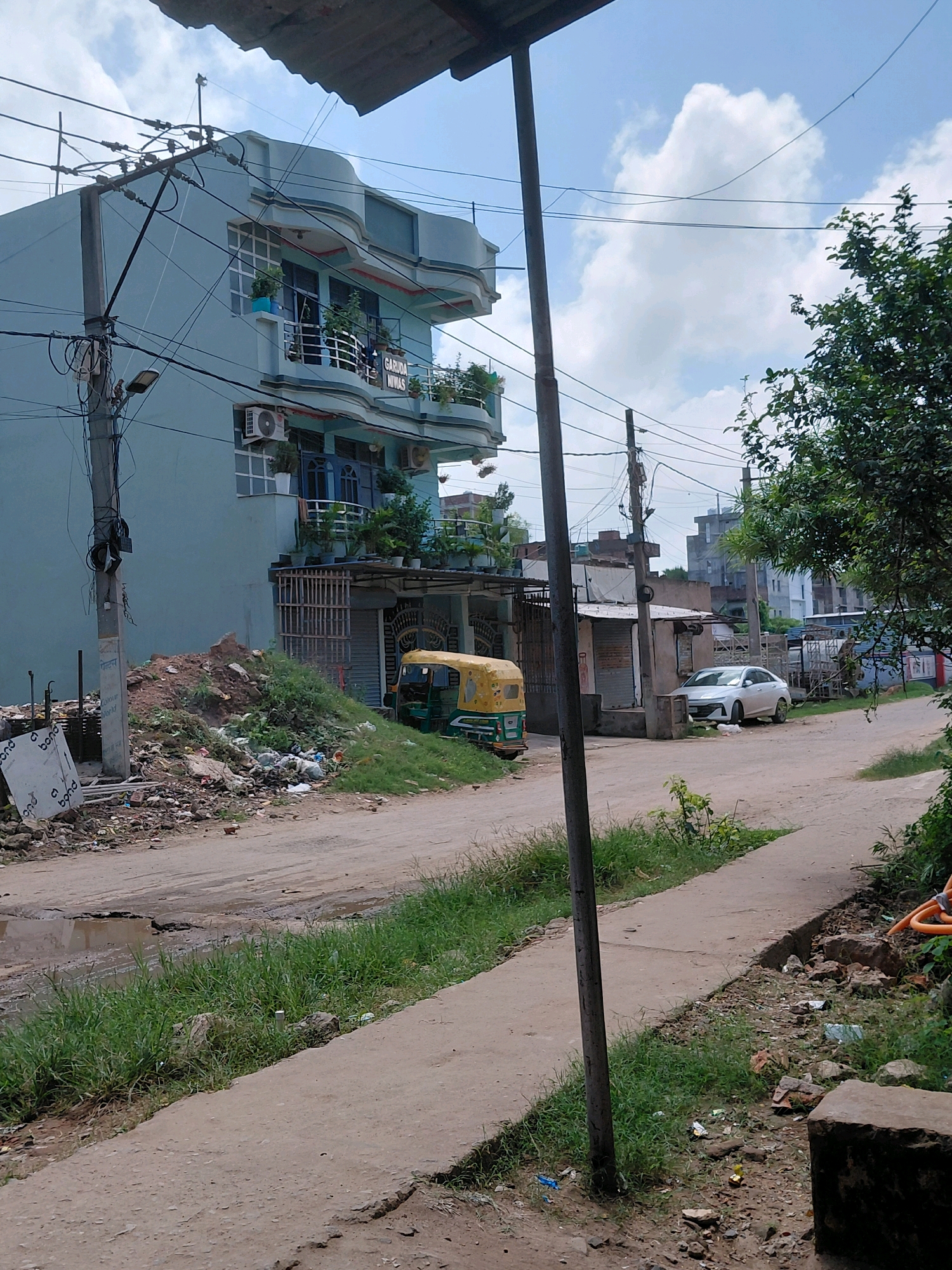 The image captures a street scene in Patna, India, characterized by a light blue, multi-story building on the left. The building features balconies with railings and plants, as well as visible air conditioning units and windows. A prominent utility pole with numerous wires runs along the left side of the image, adjacent to a pile of dirt and debris, including scattered trash and what appears to be construction materials. A yellow and green auto-rickshaw is parked near the building's entrance, which is covered by a metal grate.
Further down the street, a white sedan is parked in front of a small, roadside structure. The road itself is unpaved and appears to have some puddles or uneven surfaces. The right side of the image is dominated by a large, leafy green tree. The sky above is partly cloudy. The overall impression is of a developing urban or semi-urban area with elements of both residential and commercial activity, alongside visible signs of disarray and ongoing construction or neglect.