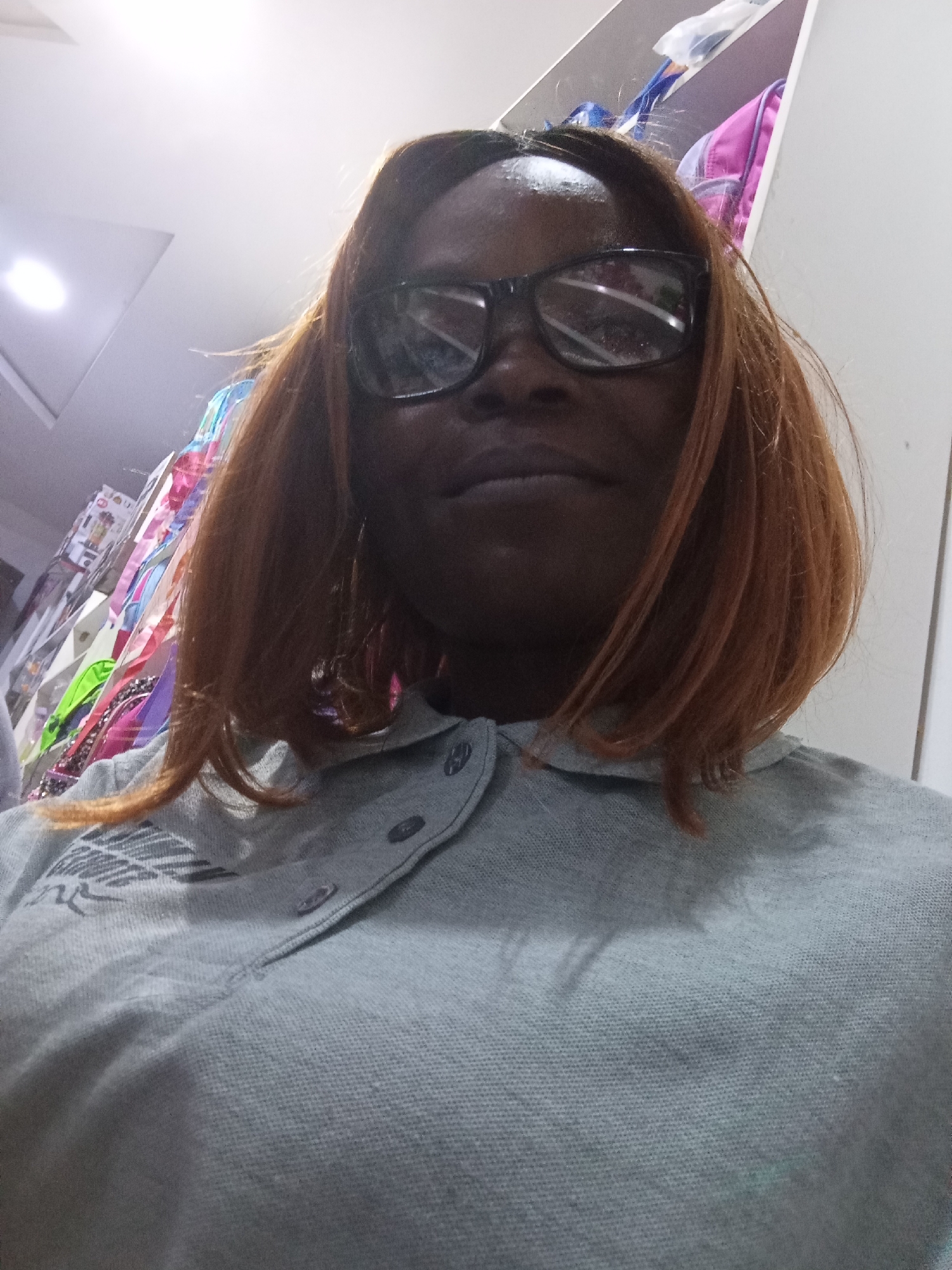 A woman wearing glasses and a grey collared shirt is pictured from a low angle, looking upwards. Her hair is shoulder-length and auburn. The background shows a brightly lit ceiling, shelves with various colorful items, and a hint of a white wall. The environment appears to be an indoor retail space, possibly a store, with merchandise displayed on shelves.