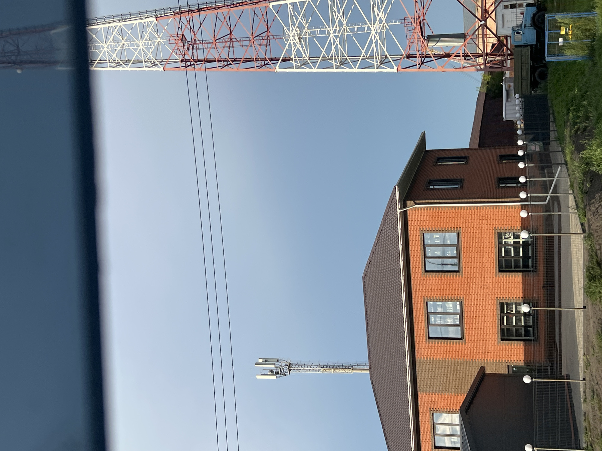 The image shows a view from a vehicle, likely a car, looking towards a brick building and two tall towers against a clear blue sky. The building is red brick with a brown roof and multiple windows. To the left of the building is a smaller, dark-colored structure. In front of the building, a black metal fence with spherical lights on posts runs along a path.
Dominating the right side of the image is a large, red and white lattice tower, characteristic of a telecommunications tower. To the left, in front of the brick building, is a second, slimmer tower with antennas at the top. In the background, behind the fence and to the right of the brick building, a blue truck is parked near some white containers and a building. Overhead, three dark electrical wires stretch across the sky. The foreground shows green grass and some vegetation. The scene appears to be set in an open area, possibly an industrial or semi-rural part of Bolshoy Oyosh, Russia. There are no people visible in the image.