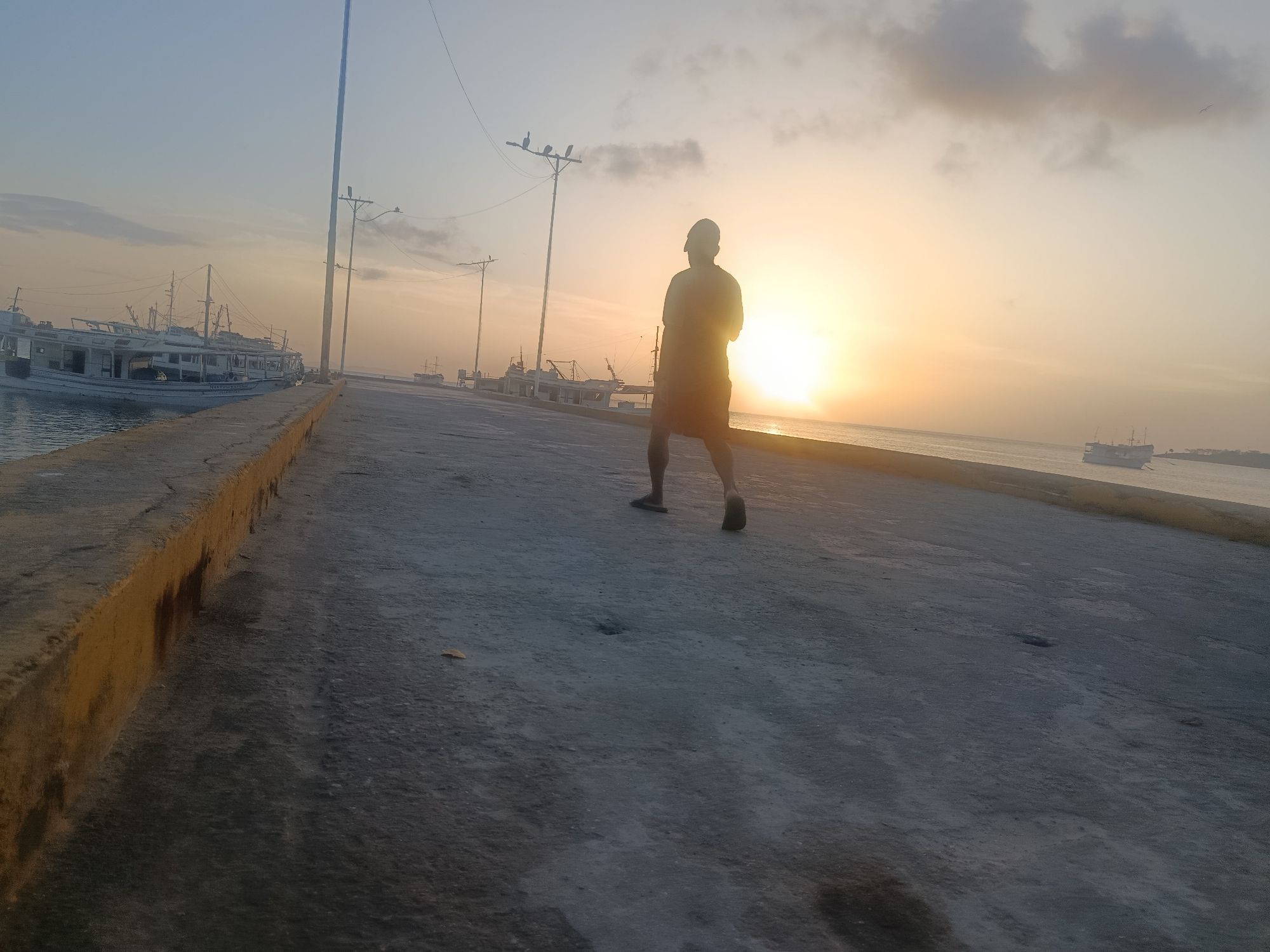 The image depicts a person walking away from the camera on a pier at sunrise in Juan Griego, Venezuela. The sun is low on the horizon, casting a warm glow over the scene. Several boats are docked in the background, suggesting this is a harbor. The pier is made of concrete and has a slightly rough texture. The person is wearing shorts and a t-shirt. The sky is a mixture of blue and orange, with some clouds visible. The overall impression is one of peace and tranquility.