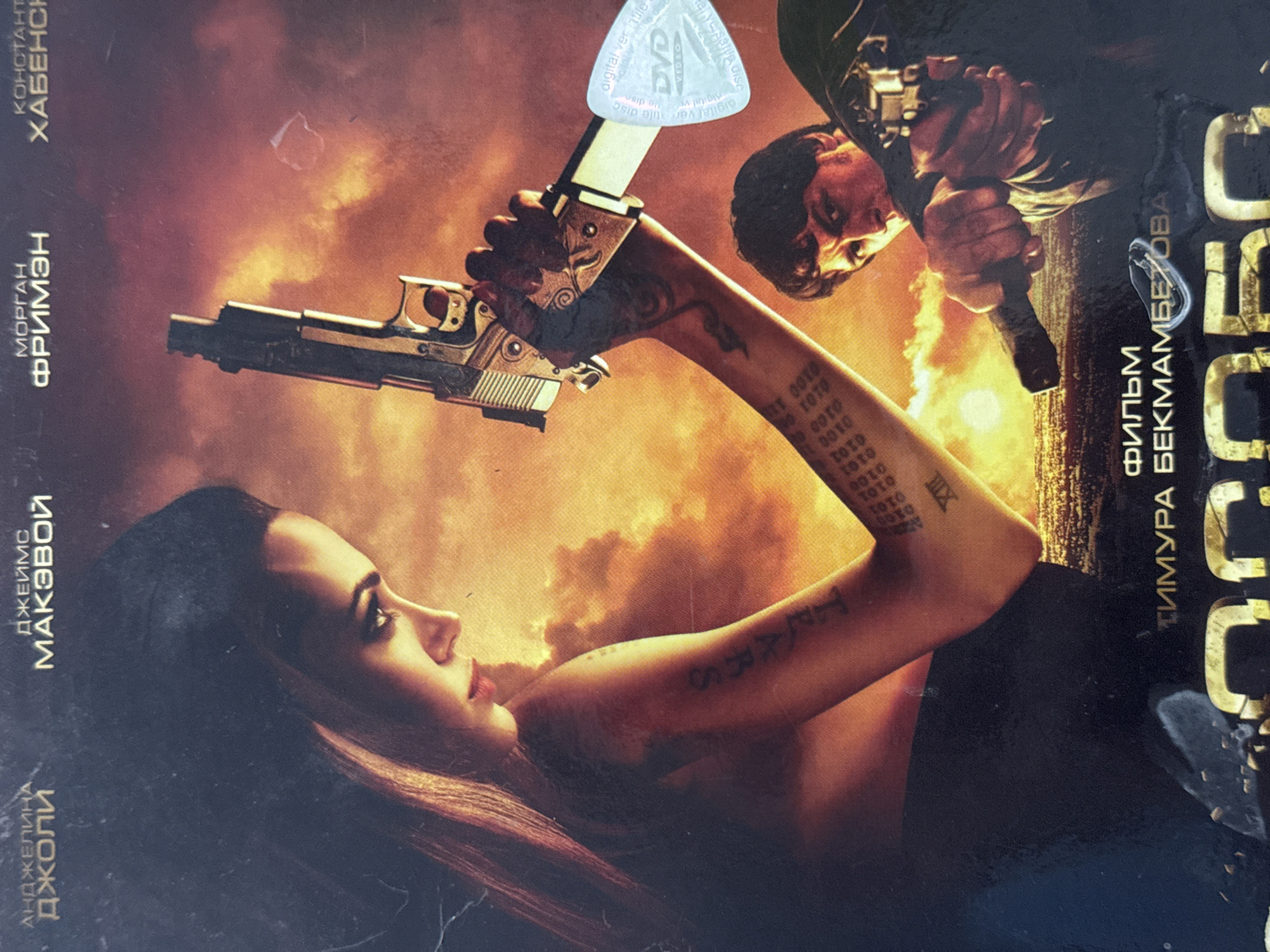 This image is a movie poster, featuring Angelina Jolie prominently. She is lying on her side, holding a silver-colored handgun above her head. Her arm has various tattoos, including "TRARS," a numerical sequence of "0101," and the Roman numeral "XIII." Another figure, a man, is partially visible in the upper right corner, also holding a gun. A "DVD Video" sticker is affixed to the poster. The background appears to be fiery and smoky. The poster also includes names of actors and the director in a Cyrillic alphabet. The words “АНДЖЕЛИНА Джоли”, “ДЖЕЙМС МАКЭВОЙ”, “МОРГАН ФРИМЭН”, and “КОНСТАНТ ХАБЕНС” are displayed. There are no obvious cues that the image was captured in Chuprino, Russia.