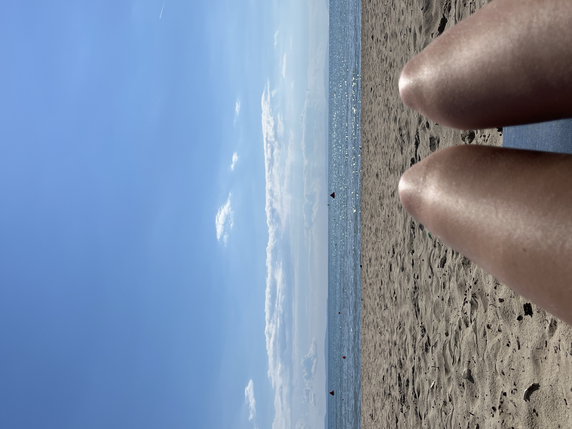 The image shows a first-person perspective of someone sitting on a beach, with their legs extending towards the foreground. The sand is a light brown color and shows the texture of footprints and small debris. In the midground, the ocean is visible, appearing calm with gentle waves and sparkling reflections from the sun. A thin, hazy line of land or islands is visible on the horizon. The sky is a clear, bright blue with scattered, fluffy white clouds, particularly in the mid-sky. There are two red buoys visible in the water, indicating a marked area. The overall environment is a sunny beach scene. The specific location is Unknown, Greece.