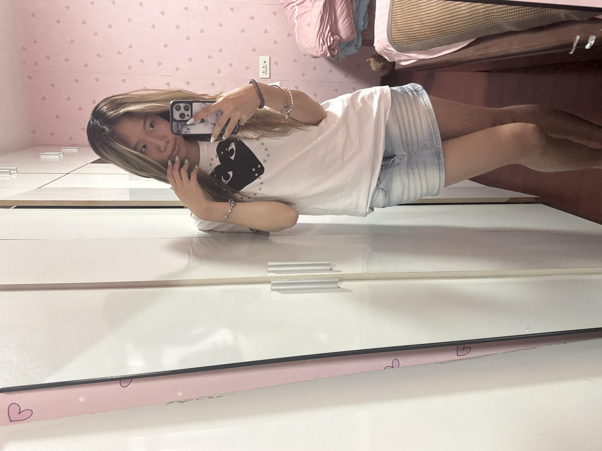 A young woman is taking a selfie in a mirror. She is wearing a white t-shirt with a black heart graphic and light blue denim shorts. She has brown hair with blonde highlights. Her phone has a clear case. The background includes a pink wallpaper with small heart designs. A bed with pink and tan blankets is visible in the upper right corner. The mirror frame is decorated with pink hearts. The location is most likely an apartment in Shapingba District, China.
