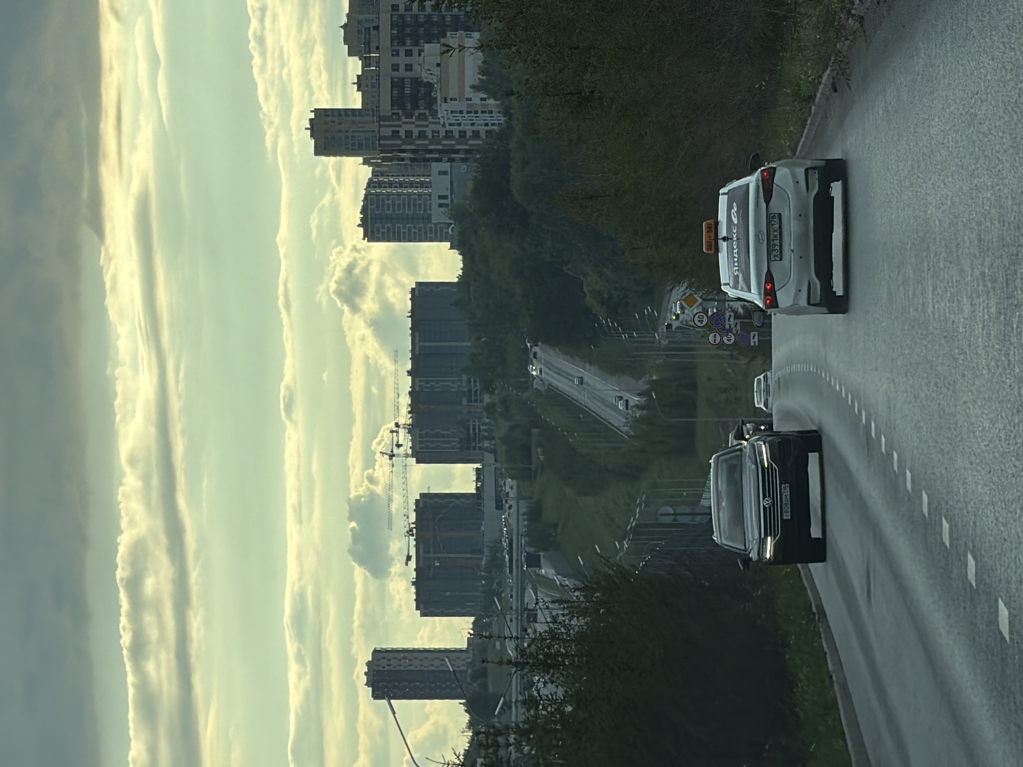 The image depicts a view from a vehicle driving on a highway, looking forward and slightly upward. The scene is characterized by a wide highway with two lanes visible in the foreground, both heading uphill. On the right lane, a white Hyundai sedan, clearly marked as a Yandex taxi, is in motion. The car has a bright orange taxi sign on its roof and Yandex Go branding on its rear. Further ahead, in the left lane, a dark-colored Volkswagen SUV is also moving uphill. Beyond the highway, a densely wooded area is visible, with the terrain rising into rolling hills. In the distance, a cityscape emerges, featuring several modern, multi-story buildings, some of which appear to be under construction, indicated by cranes. The sky above is filled with dramatic, layered clouds, tinged with the soft, diffused light of either early morning or late afternoon. The overall atmosphere is that of urban development encroaching on a natural landscape, under a moody sky.