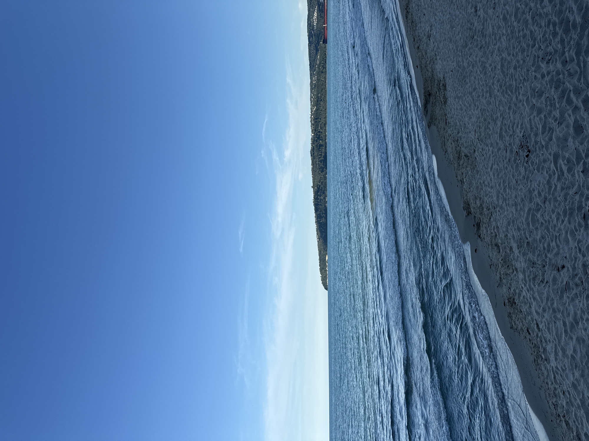 A wide shot captures a serene beach scene under a clear blue sky with a few wispy clouds. The foreground is dominated by a sandy beach, showing intricate patterns of sand ripples. The gentle turquoise water of the sea meets the shore with small waves lapping at the sand. In the distance, a lush green hillside is visible, dotted with buildings and trees, suggesting a coastal town. A large red ship or structure is also present on the right side of the frame, partially obscured by the distant landscape. The overall impression is one of natural beauty and tranquility.