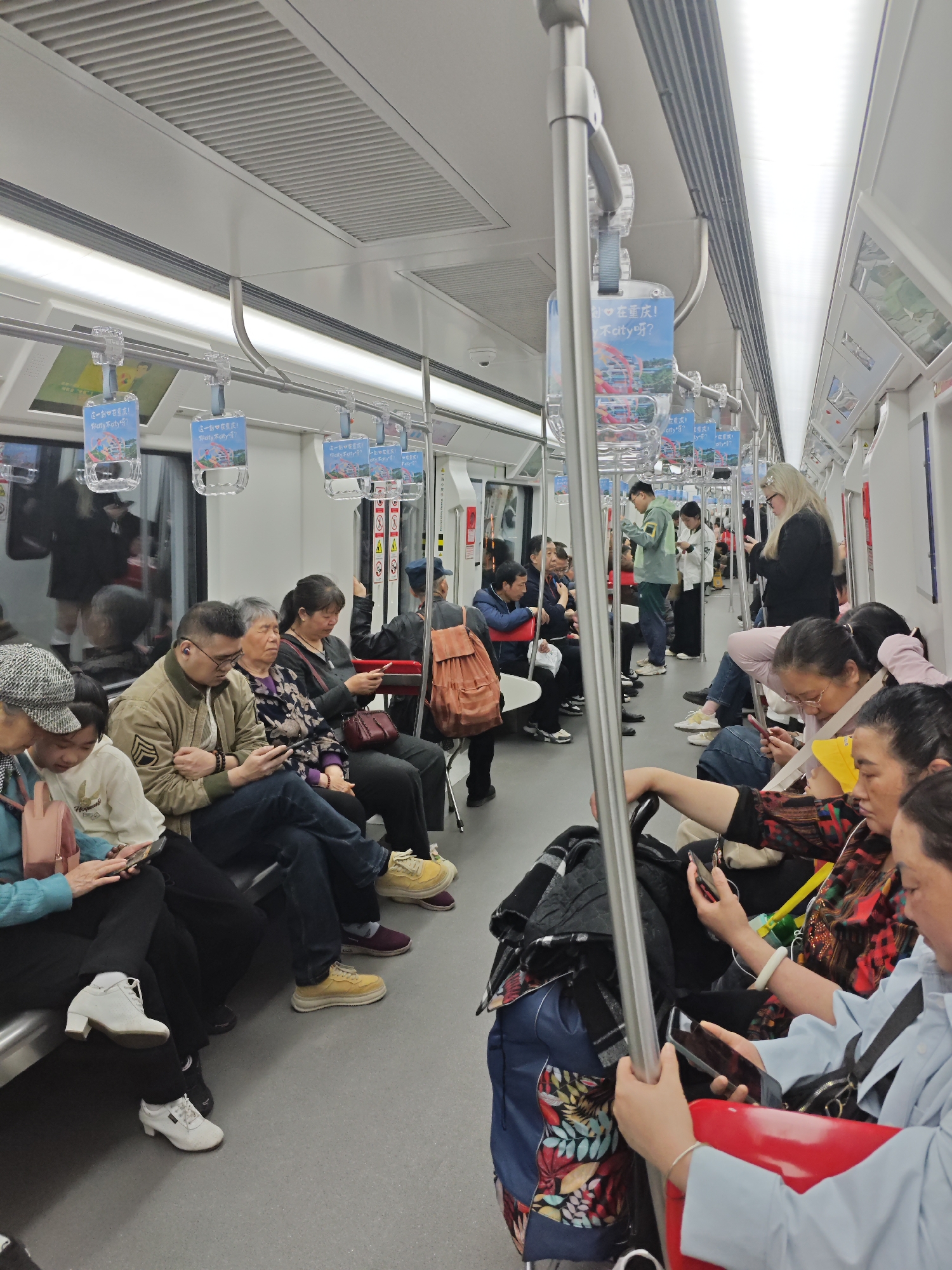 The image shows the interior of a crowded train car, likely a subway or metro, with passengers occupying most of the seats and some standing. Most of the people are Asian. Many passengers are looking at their phones, while others are seated and observing their surroundings. Overheads are advertising posters, which include the Chinese text "这一刻在重庆!” “你atyaty吗?" and “不city呀?" The posters indicate the location is Chongqing, China. The train car has a neutral color scheme with white walls and a grey floor. The interior suggests the train is modern. The photograph was likely taken in Shapingba District, China.
