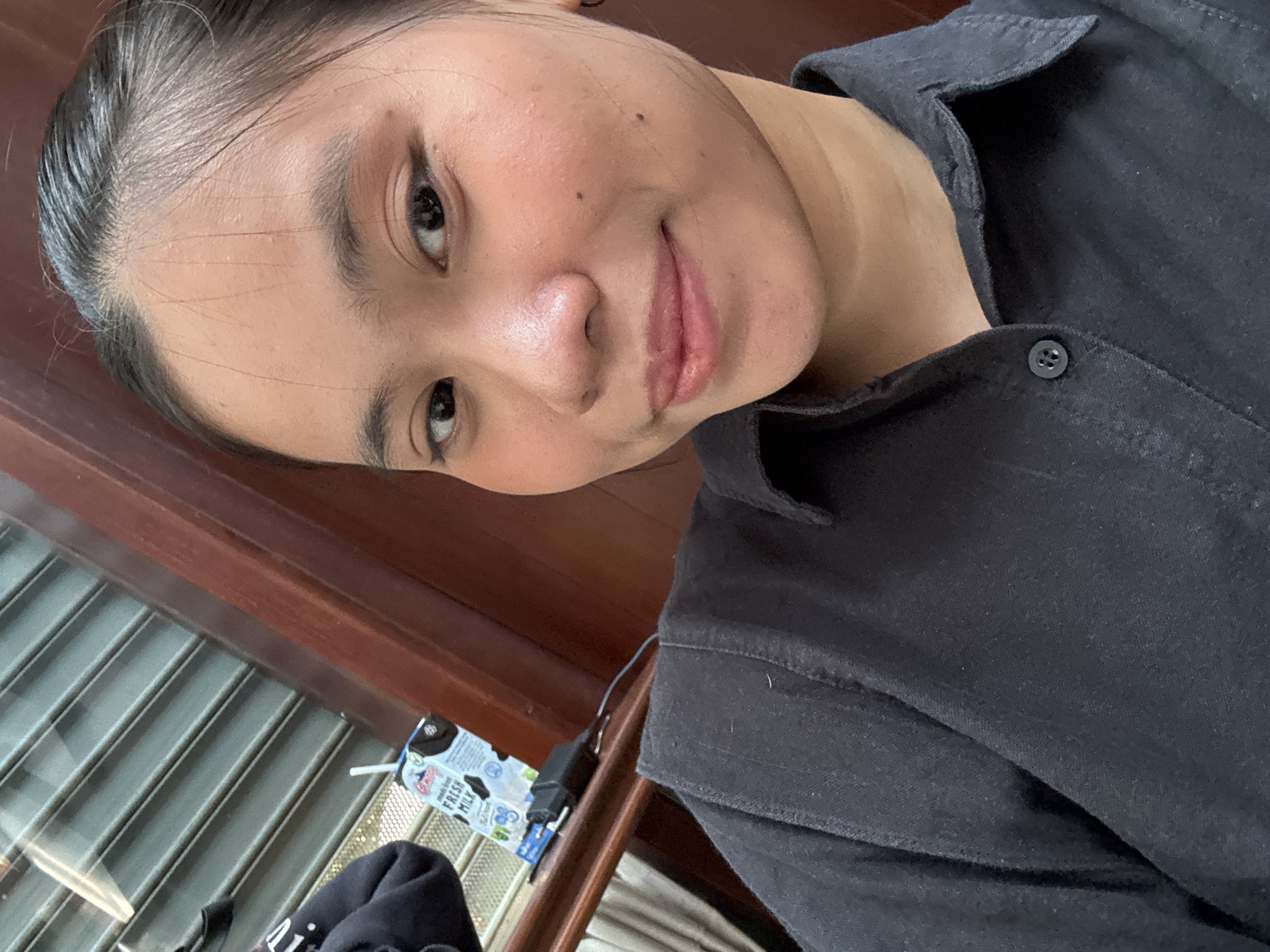 A close-up, selfie-style photograph captures a young East Asian woman with dark hair pulled back from her face. She has dark eyes, a light brown complexion, and is wearing a black collared shirt. Her makeup includes subtle eyeliner and lip color. She is positioned in front of a wooden surface and what appears to be a window with horizontal blinds. To her left, a carton of "Fresh Milk" with a straw is visible, along with a black electrical plug. The overall lighting suggests an indoor setting, possibly in a residential or commercial space. The woman is looking directly at the camera with a faint smile. There is no clear indication of the specific location within South Jakarta, Indonesia, beyond the possibility of an urban environment.