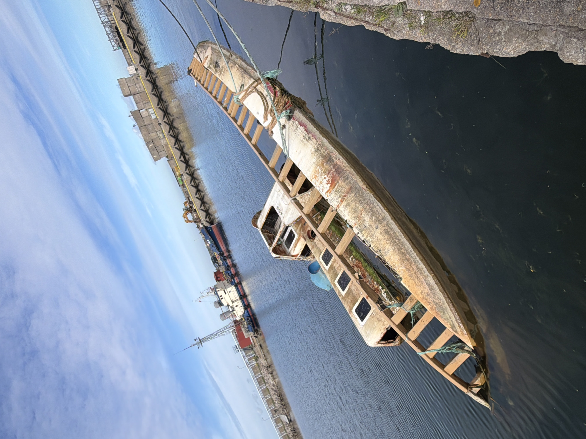 This is a photograph taken in the city of Viimsi, Estonia, of a harbor area. In the foreground, a partially submerged, derelict vessel with a wooden structure on top lies in the dark blue water. The vessel is white with rust-colored patches and is secured by ropes to what appears to be a concrete pier on the right side of the frame. Behind this derelict vessel, a larger, operational ship with a white superstructure and a blue hull is docked. Further in the background, a concrete pier is visible, lined with yellow buoys and industrial equipment, including what appears to be an excavator. Beyond this, a large ship with a tall mast and antenna is moored. The sky above is a bright blue with scattered white clouds, and the sun appears to be shining, casting reflections on the water. The overall scene depicts a working harbor with both active and abandoned vessels.