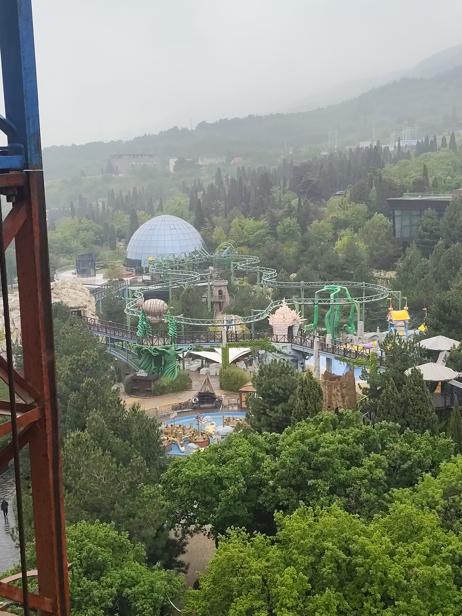 The image depicts an overview of a portion of a theme park located in Симеизский поселковый совет, Ukraine. A prominent feature is a geodesic dome, possibly a planetarium or a themed attraction, situated near a green-painted roller coaster. The coaster snakes through the park's landscape, which is heavily vegetated with mature trees. Several themed structures and attractions are visible, including what appears to be a water ride with boats. An observation tower provides an elevated vantage point. In the lower left corner, a person can be seen walking along a paved path. The weather appears overcast, with a hazy or foggy background.