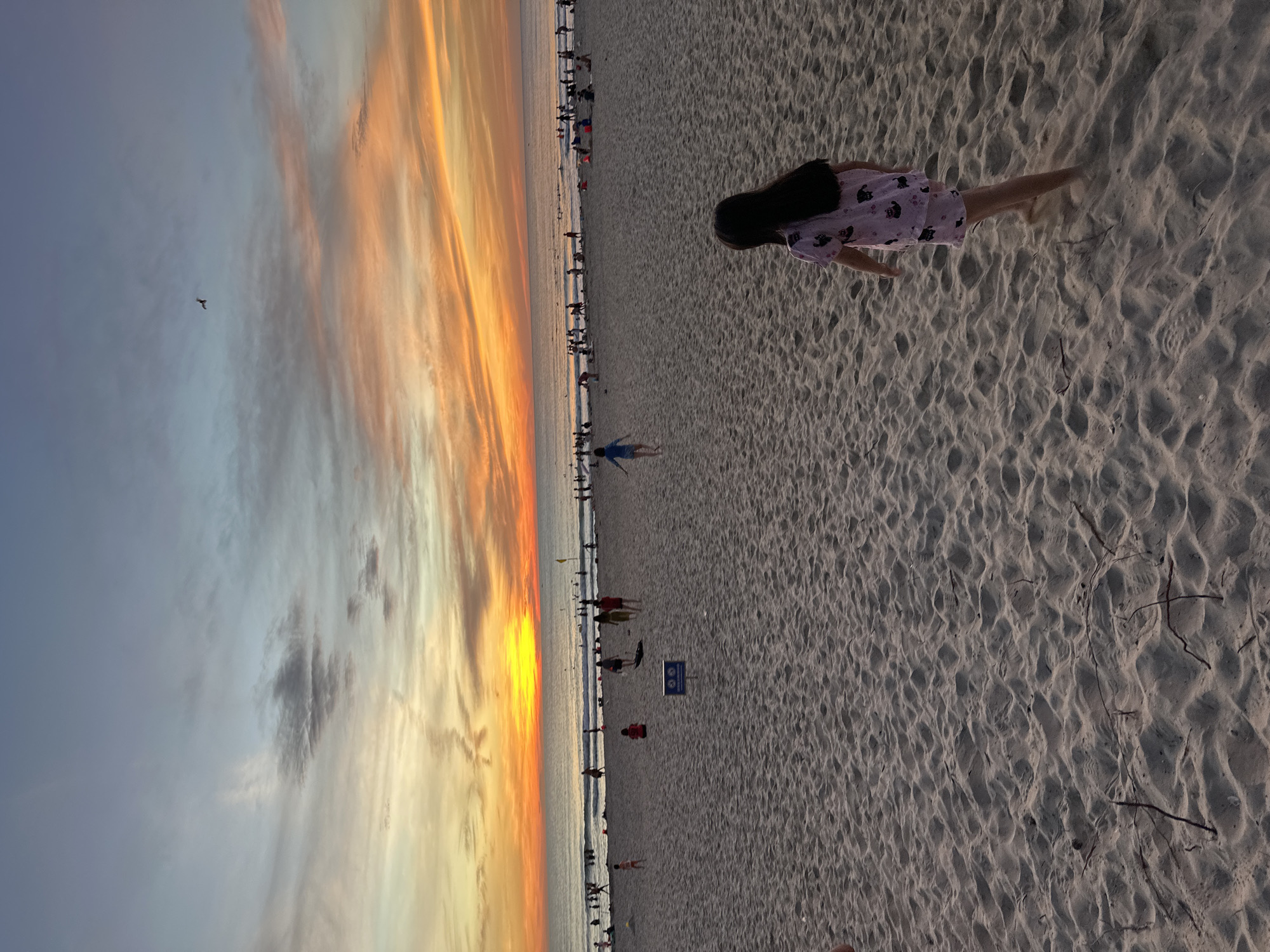 The image depicts a beach scene at sunset. The sky is filled with vibrant colors, ranging from deep orange and yellow near the horizon to soft blues and grays higher up, with wispy clouds scattered throughout. A single bird is visible in flight in the upper right portion of the sky. The ocean stretches across the background, with gentle waves lapping at the shore. Numerous people are present on the beach and in the water, engaged in various activities such as swimming, walking, and playing. In the foreground, a young girl with long dark hair, wearing a pink outfit with a pattern of black cats, is walking away from the camera on the sandy beach. The sand is rippled, indicating the movement of water or wind. A blue sign with white symbols is partially visible in the middle ground. The overall impression is that of a lively and picturesque beach at dusk. The location is identified as Đà Nẵng, Vietnam.