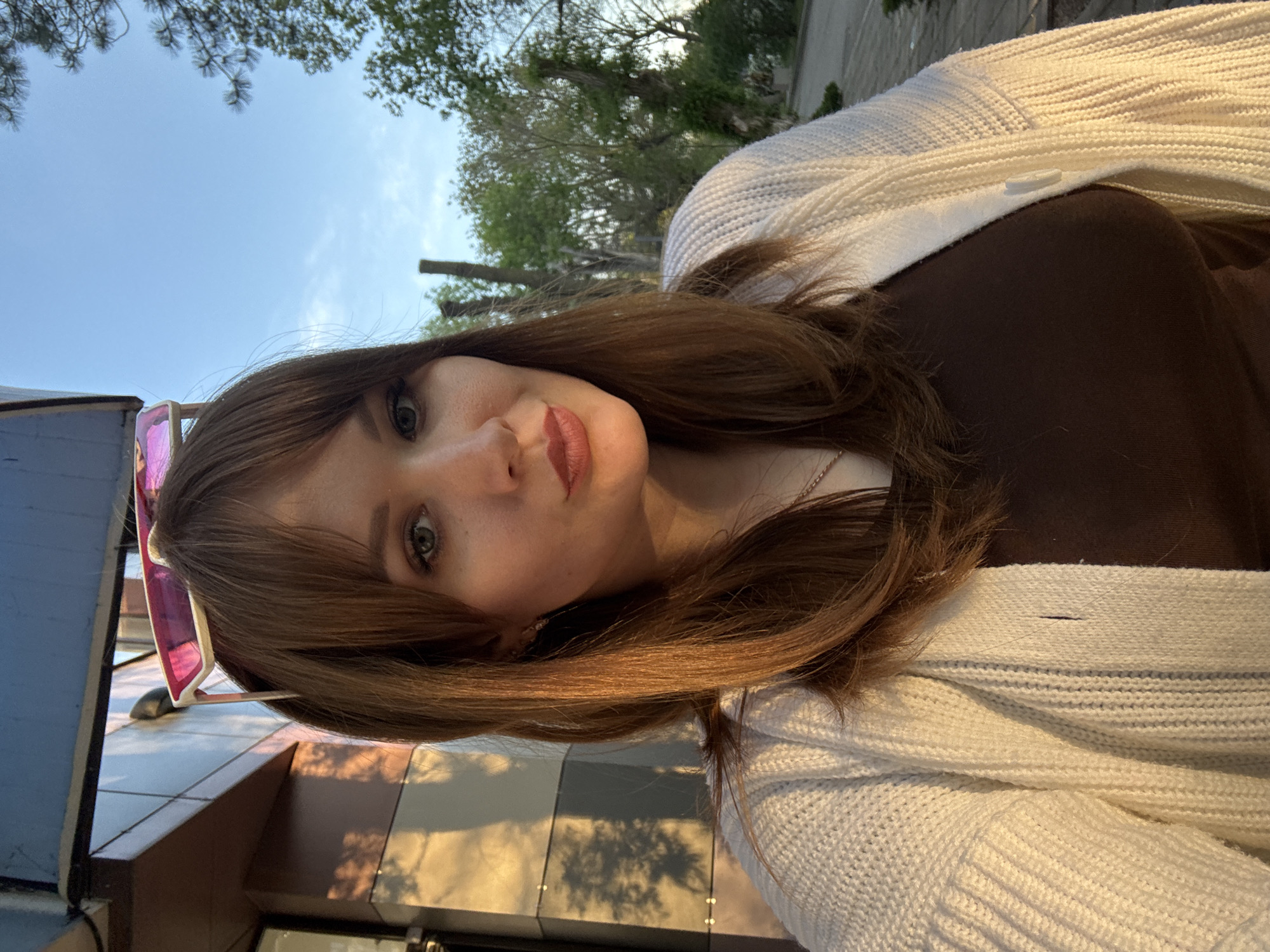 A woman is posing for a selfie outdoors in Yevpatoriya, Ukraine. She has medium-length auburn hair with bangs and is wearing a brown top and a white knit cardigan. Pink-framed sunglasses are perched on top of her head. In the background, there are trees and a building, partially obscured by shadows. The sky is visible in the upper left corner, suggesting it is daytime.