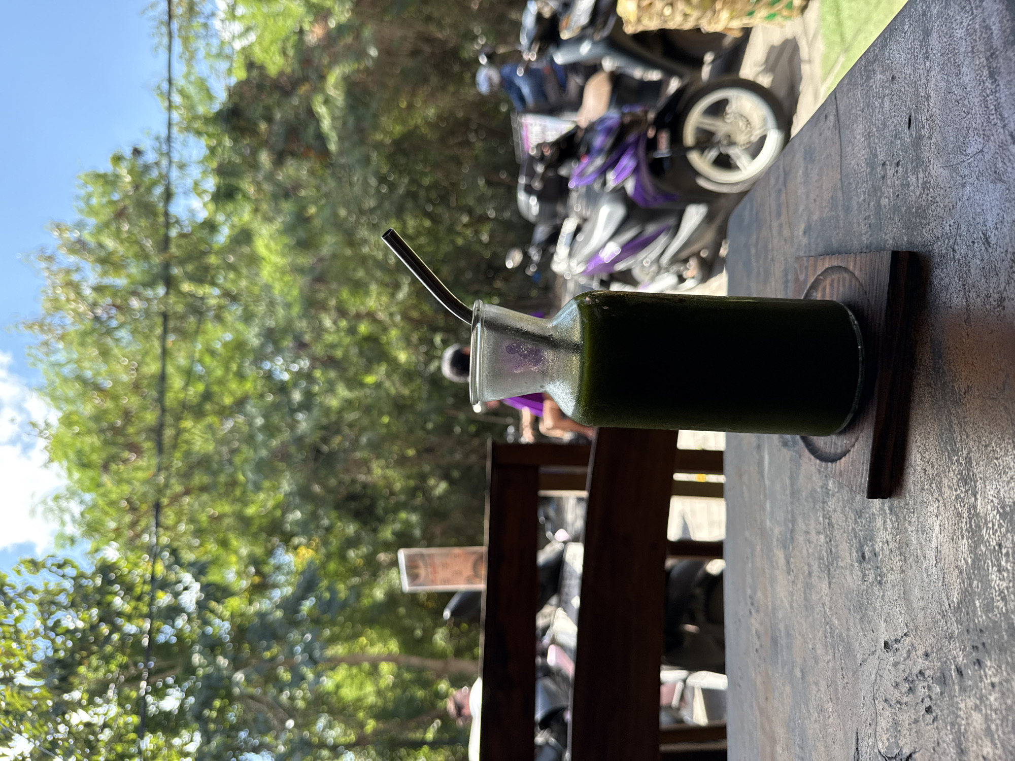 A glass bottle containing a dark green beverage, with a silver metal straw inserted, is the central focus of the image. The bottle rests on a round, carved wooden coaster, which in turn sits on a rustic, textured dark wooden table. The table surface shows natural grain and some wear.
In the background, lush green tree foliage dominates the left side of the frame, suggesting an outdoor setting. Above the trees, a bright blue sky with a few faint white clouds is visible. Beyond the table and through the trees, several parked motorbikes are partially visible; some have purple and black accents. The scene is brightly illuminated by natural sunlight, indicating daytime. The composition suggests a relaxed atmosphere, possibly at an outdoor cafe or eatery in Kuta Selatan, Indonesia. There is no visible text in the image.