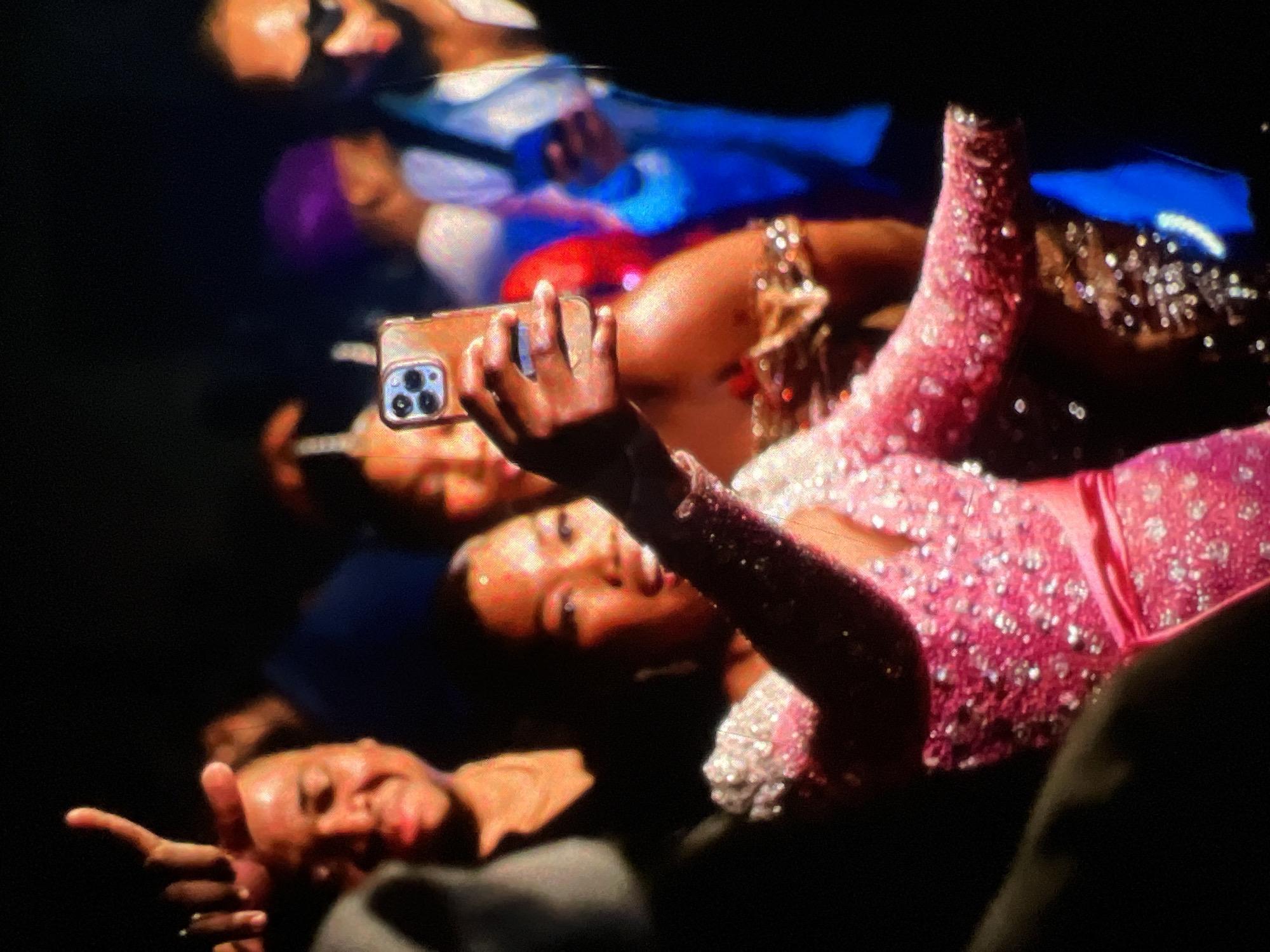 The image depicts a group of people in a dimly lit indoor setting, possibly a party or event, with a focus on a woman in the foreground holding up a smartphone to take a selfie. She is wearing a sparkling pink dress with a V-neckline and a matching pink belt. To her left, a man is partially visible, smiling and holding up two fingers, possibly in a gesture of victory or peace. In the background, other individuals are present, including a person wearing a purple headscarf and another wearing sunglasses. The overall ambiance suggests a lively social gathering. The exact location in Lagos, Nigeria, cannot be determined from the image alone.