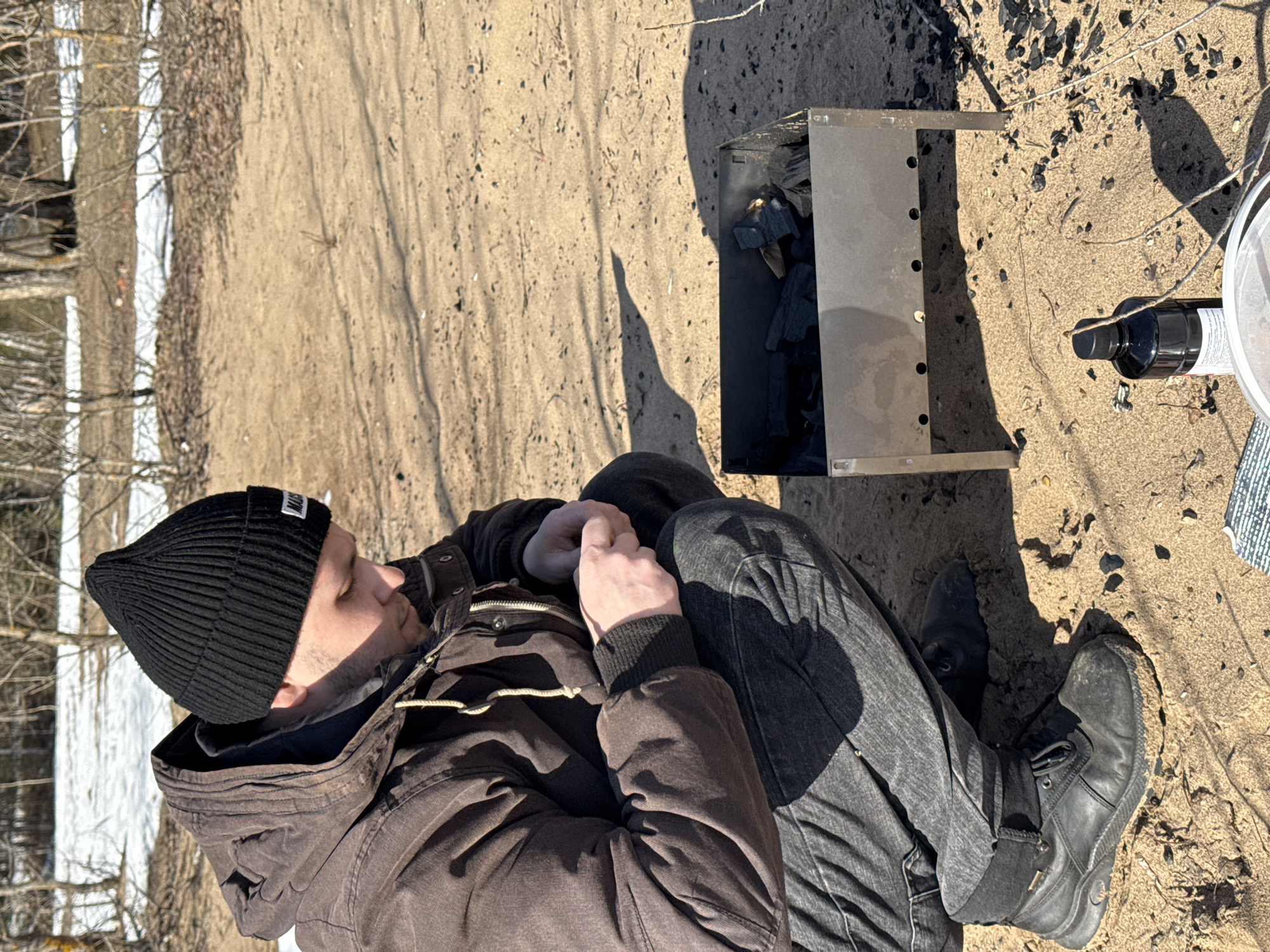 A ground-level, angled photograph depicts a person sitting on the ground next to a metal grill filled with charcoal, seemingly outdoors in an area with sandy ground. The person is wearing a black beanie, a brown jacket, and dark jeans with black boots. They are sitting with their knees bent and their hands clasped. To the person's right is a small, square, metal grill containing pieces of charcoal. Next to the grill is a black bottle and a white bucket or container with a striped towel nearby. The ground is sandy and uneven. In the background to the left, there are trees and patches of snow, suggesting it is early spring. The location is not specified.