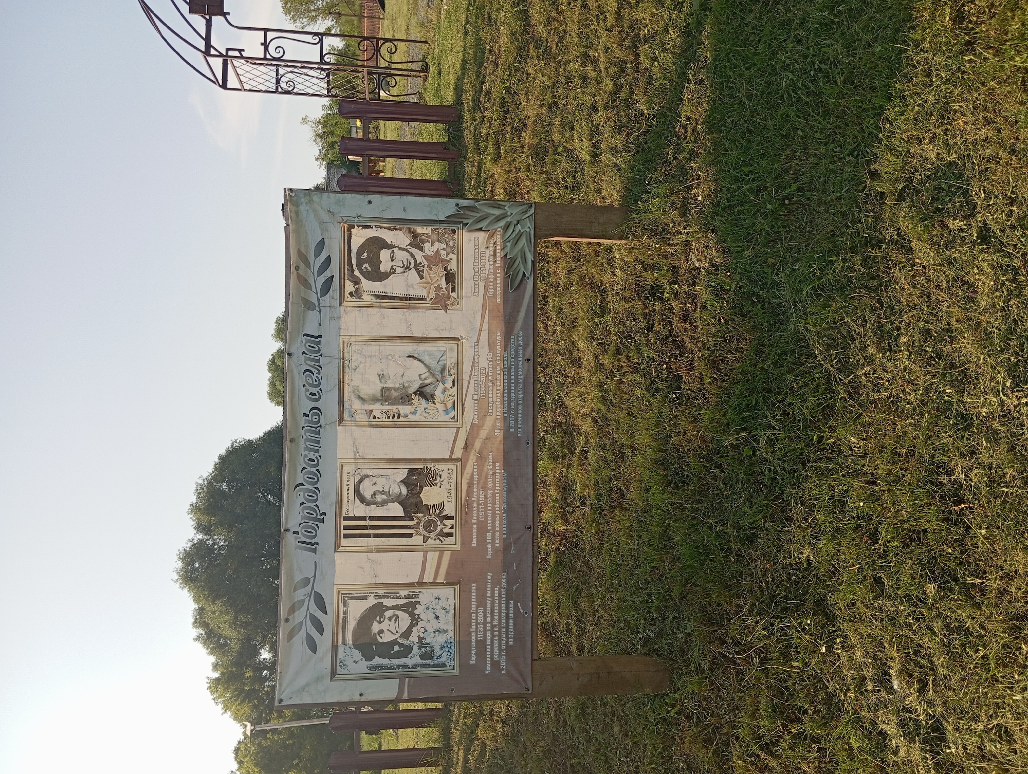 This is a full-frame, eye-level, outdoor shot of a memorial display board in the village of NovoKopylövo, Russia. The board, mounted on two wooden posts, features the title "Гордость села" (Pride of the Village) and displays four portraits with accompanying text. The portraits are of individuals, each in a framed section. The environment is a grassy area with trees and a metal archway visible in the background. The lighting suggests it is daytime, possibly morning or late afternoon due to the long shadows.
