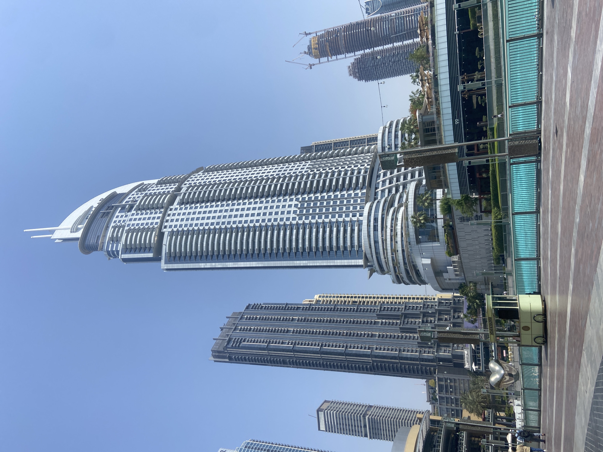 The image depicts a cityscape in Dubai, United Arab Emirates, under a clear blue sky. Dominating the view is the Address Downtown hotel, a curved high-rise building with numerous balconies, its glass facade reflecting the sky. A second, rectangular high-rise stands partially obscured behind it.
In the foreground, a small, light-colored cart with wheels sits on a brick-paved walkway. A modern sculpture of interlocking, reflective shapes is positioned nearby. Behind the cart, landscaping elements, including greenery and palm trees, are visible along a multi-level structure with blue-tinted glass railings.
In the distance, other skyscrapers are under construction, denoted by the presence of cranes atop the unfinished buildings.