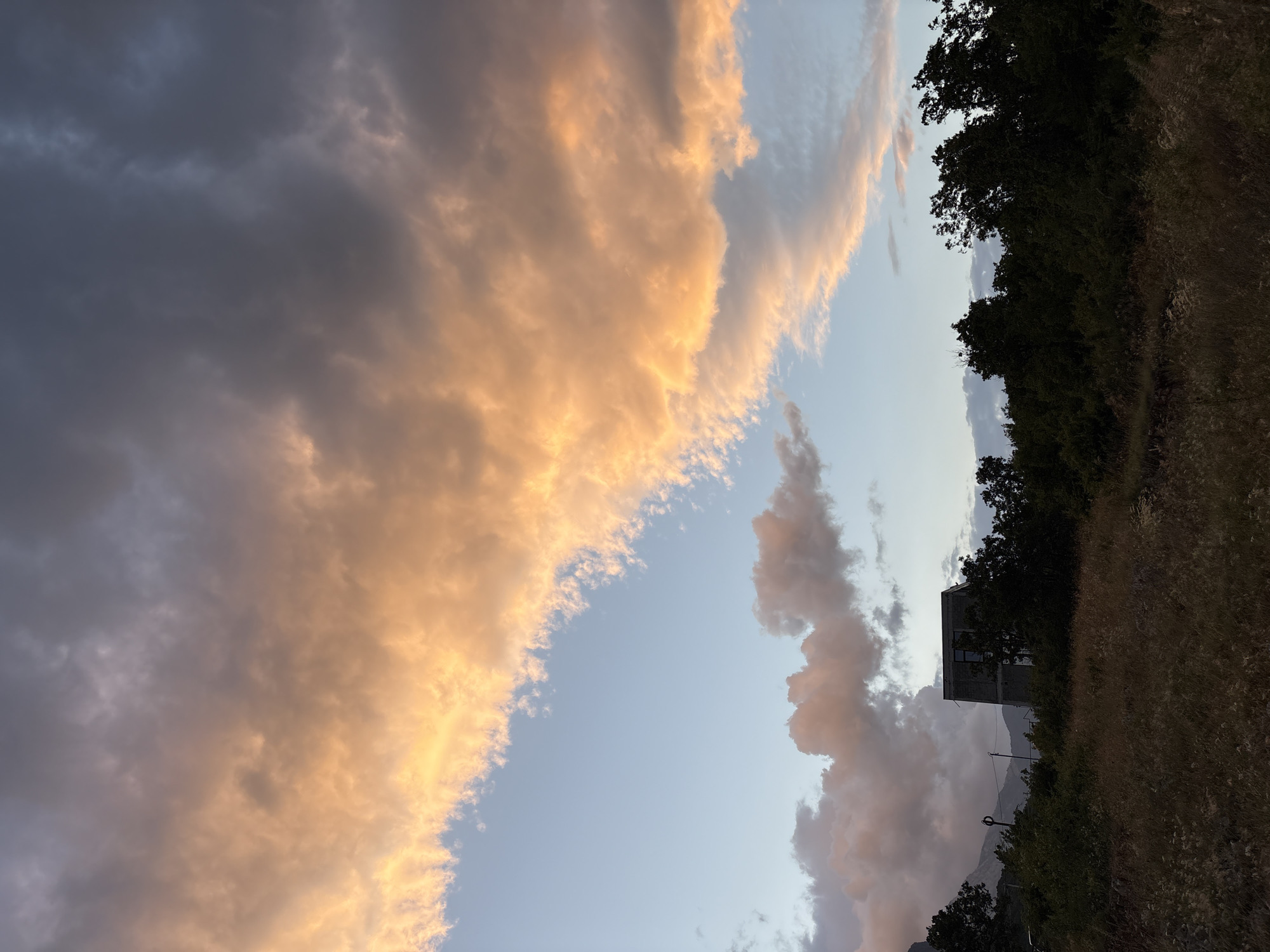 The image captures a twilight scene in Malyi Mayak, Ukraine. The sky is dominated by dramatic clouds, with a band of vibrant orange and pink hues illuminated by the setting sun stretching across the upper portion of the frame. These colorful clouds contrast with darker, brooding clouds in the upper right and softer, pinkish clouds lower in the sky.
Below the sky, a dark silhouette of trees forms a dense line across the middle ground. Peeking through the trees, a modern, multi-story building with a stark, grey facade is visible. The building appears to have several windows. In the foreground, dry, golden grass and scrub cover the ground, suggesting a rural or undeveloped area. In the far background, the faint outline of mountains can be seen. The overall impression is one of a serene yet dramatic natural landscape at dusk, with a solitary man-made structure integrated into the environment. There are no people or animals depicted in the image.