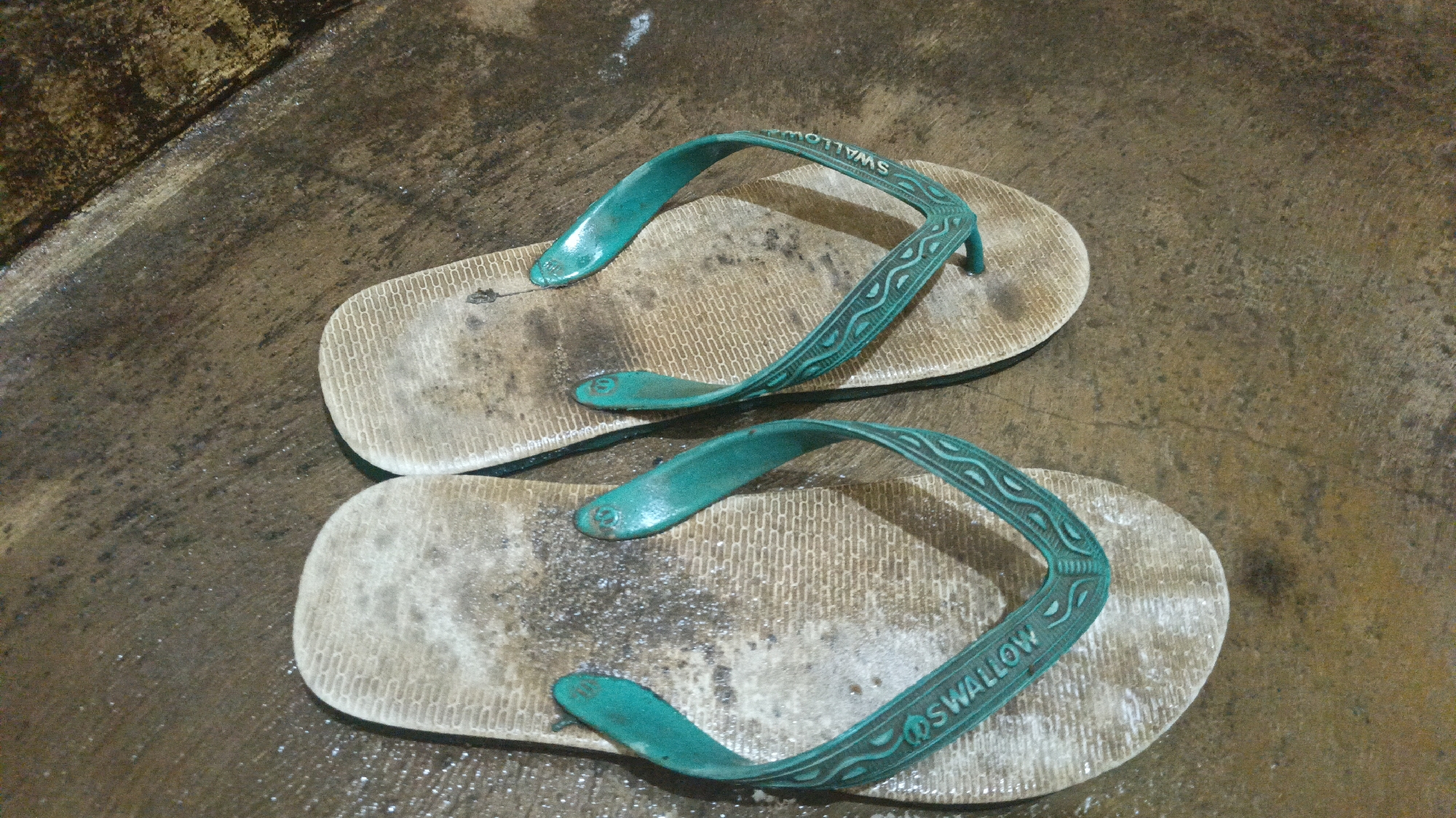 A pair of worn, teal-colored flip-flops with a white footbed and raised pattern rests on a wet, textured concrete surface. The left flip-flop is positioned slightly above and to the left of the right flip-flop. Both flip-flops show signs of wear, with dirt and scuff marks visible on the footbeds and straps. The word "SWALLOW" is embossed on the straps of both flip-flops. The concrete surface appears to be outdoors, with a darker, possibly stained area to the upper left of the frame. The scene is lit from above, casting faint shadows. This image was taken in Bayeman, Indonesia.