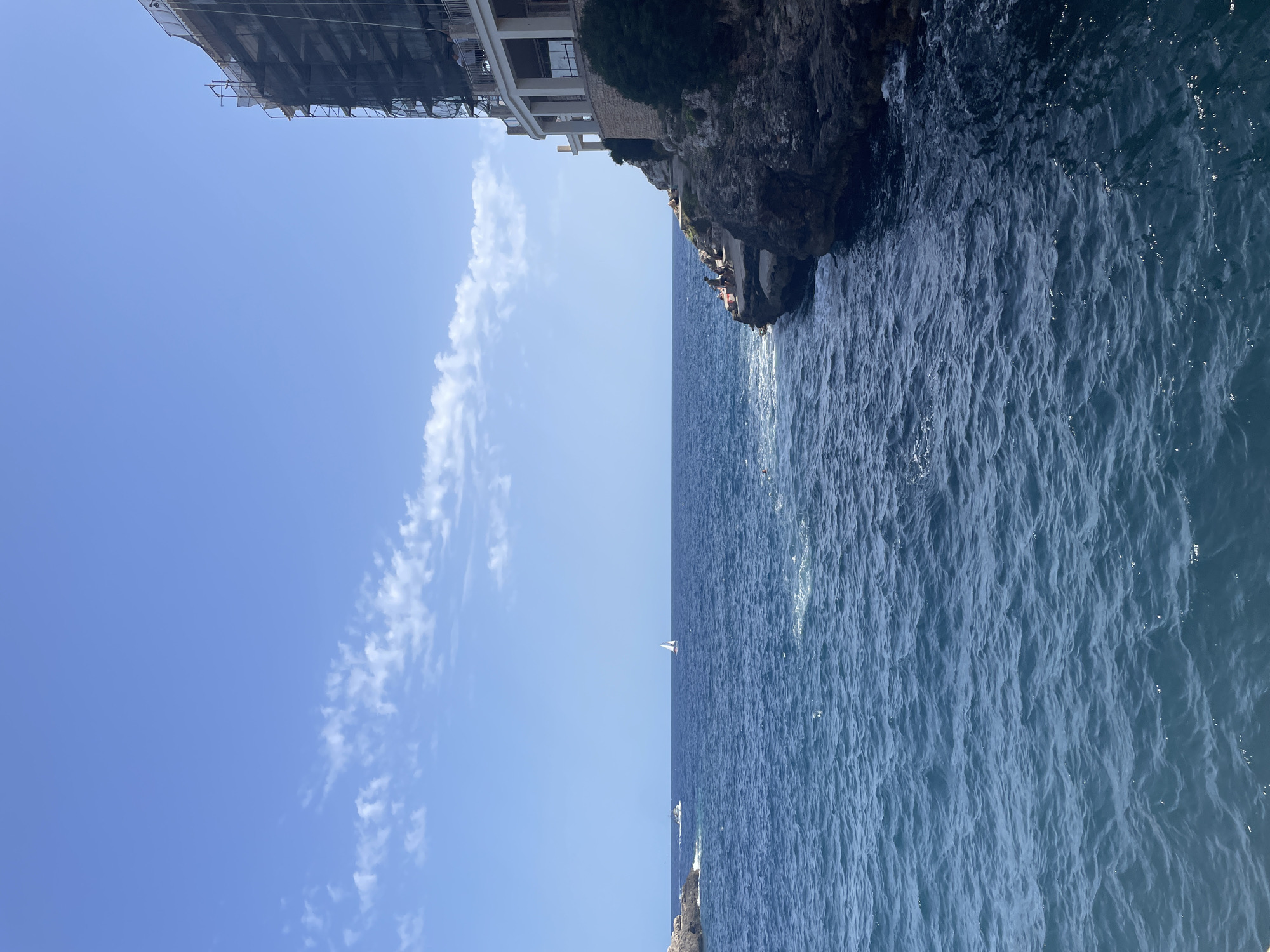 A wide shot shows a bright blue sky with sparse white clouds over a deep blue sea. On the right side of the frame, a cliff juts out into the water, topped by a multi-story building under construction, its exterior covered in scaffolding and netting. Several people are visible on the cliffside, some sitting and others near the water's edge. The sea is calm with gentle waves, and a sailboat is visible in the distance. To the left, a small rocky island is present, with a boat nearby. The scene appears to be set along the coast of Santanyí, Spain.