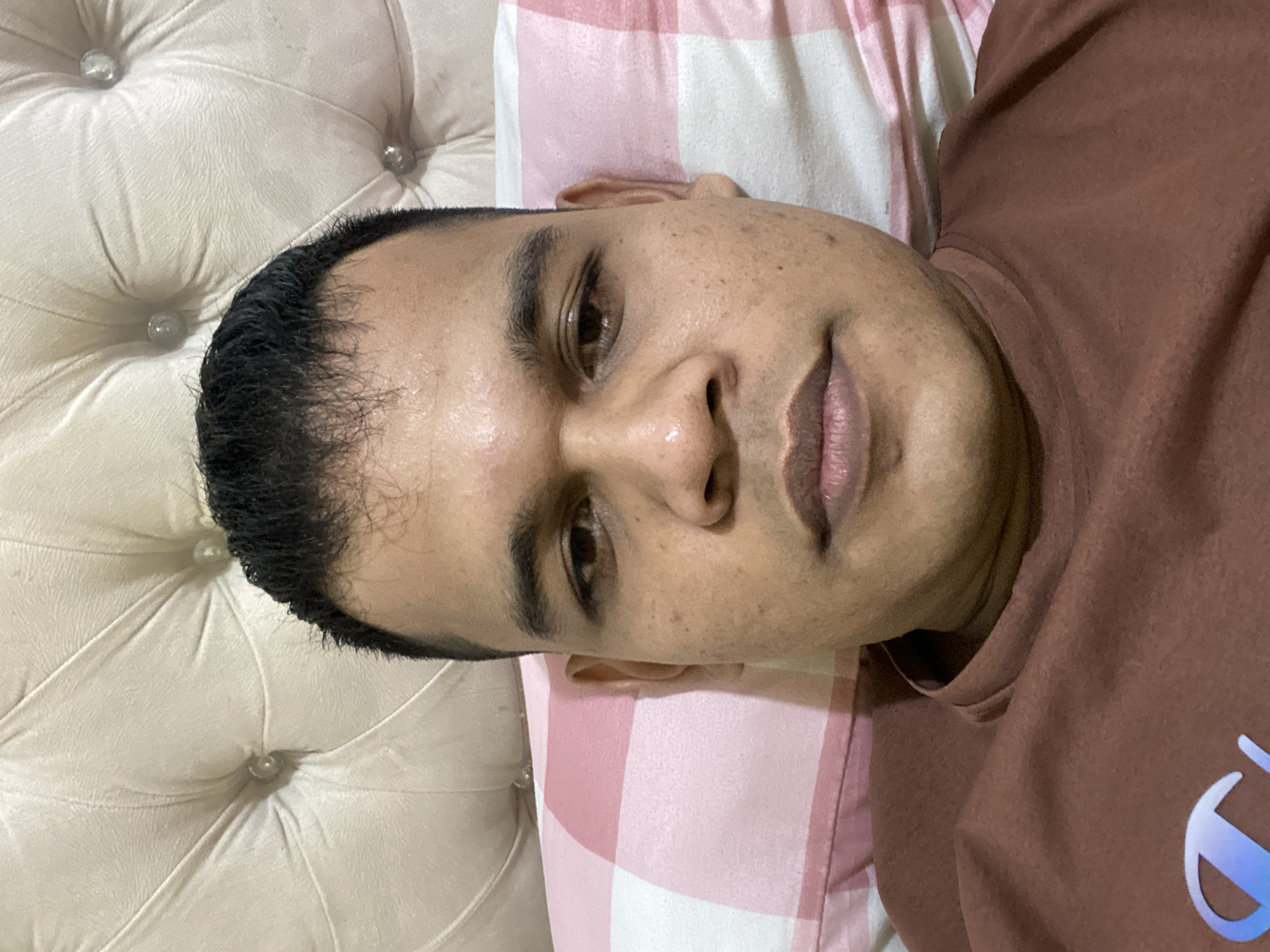 A close-up, head-on selfie of a young man lying down. He is wearing a brown t-shirt with a light blue logo. The background consists of a tufted cream-colored headboard and a pink and white checkered pillow. The man's skin appears to have some texture and light blemishes. The image is taken in Lambaro Kueh, Indonesia.