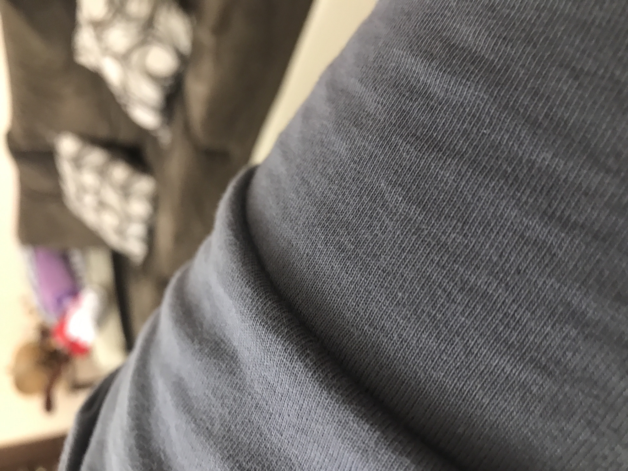 The image shows a close-up of gray fabric, likely a t-shirt or similar garment. In the background, out of focus, is a piece of furniture with patterned cushions. There are also vague shapes of what appears to be objects, possibly toys or clothing, situated further in the background. There is no evidence to suggest the location of the image is Yerevan, Armenia.