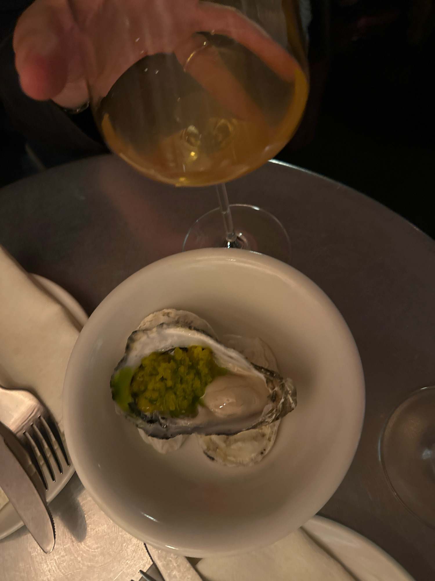 An overhead, close-up shot captures a single oyster served in a white ceramic bowl. The oyster is topped with a vibrant green relish or sauce and appears to be resting on a bed of crushed ice or rock salt, though this is not clearly visible. The bowl is placed on a metallic surface, possibly a table, which reflects the ambient light. To the left of the bowl, a fork and knife are visible, suggesting a dining setting. In the upper portion of the image, a hand is holding a wine glass filled with a pale yellow liquid, possibly white wine, with the hand partially obscuring the rim of the glass. The background is dark and out of focus, providing little environmental context. The image does not provide any discernible clues to suggest a location in Madrid, Spain.