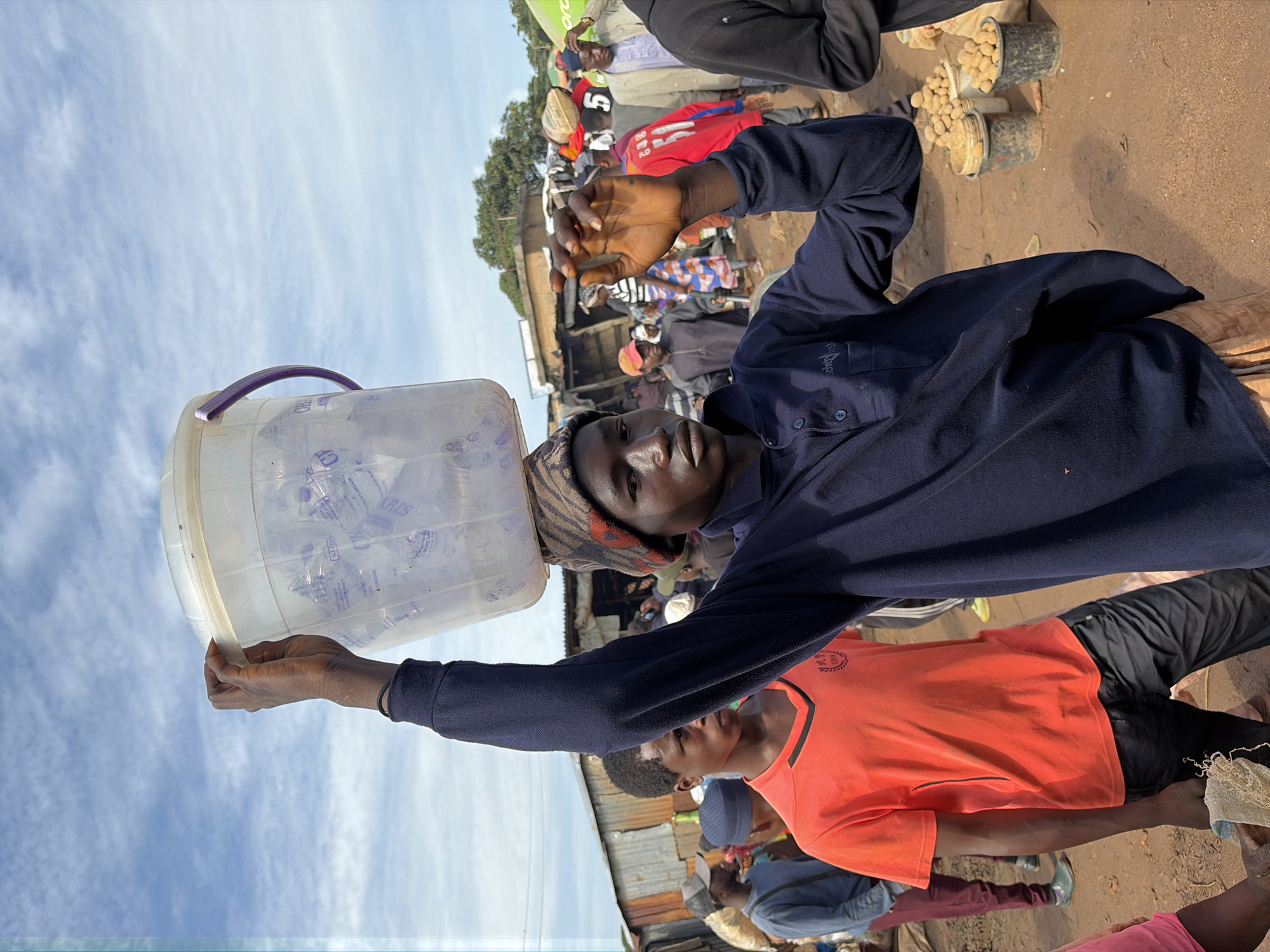 In Gura Top, Nigeria, a young Black male, wearing a dark blue long-sleeved shirt, a patterned beanie, and beige pants, balances a clear plastic bucket filled with bottled water on his head. He is walking through a bustling outdoor market with a sandy ground. In front of him, a young Black male in an orange t-shirt and black pants looks towards the camera. Behind them, a diverse crowd of people are visible, some browsing goods and others walking. Stalls and structures with corrugated metal roofs line the background. Buckets filled with potatoes are visible to the right of the main subject. The sky above is partly cloudy.