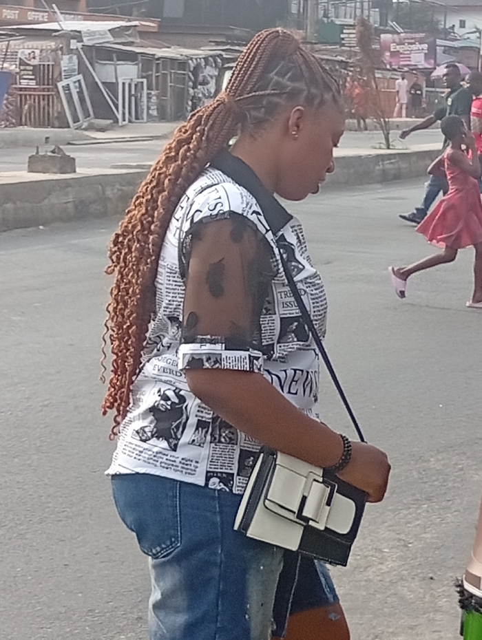 A woman stands on a street, looking down. She wears blue jean shorts, a white newspaper-print top with sheer sleeves, and has long, brown, braided hair pulled into a high ponytail. A black and white purse hangs from her shoulder. In the background are buildings, pedestrians, including a young girl in a red dress, and what appears to be a "Post Office" sign. The day is bright with diffuse lighting.